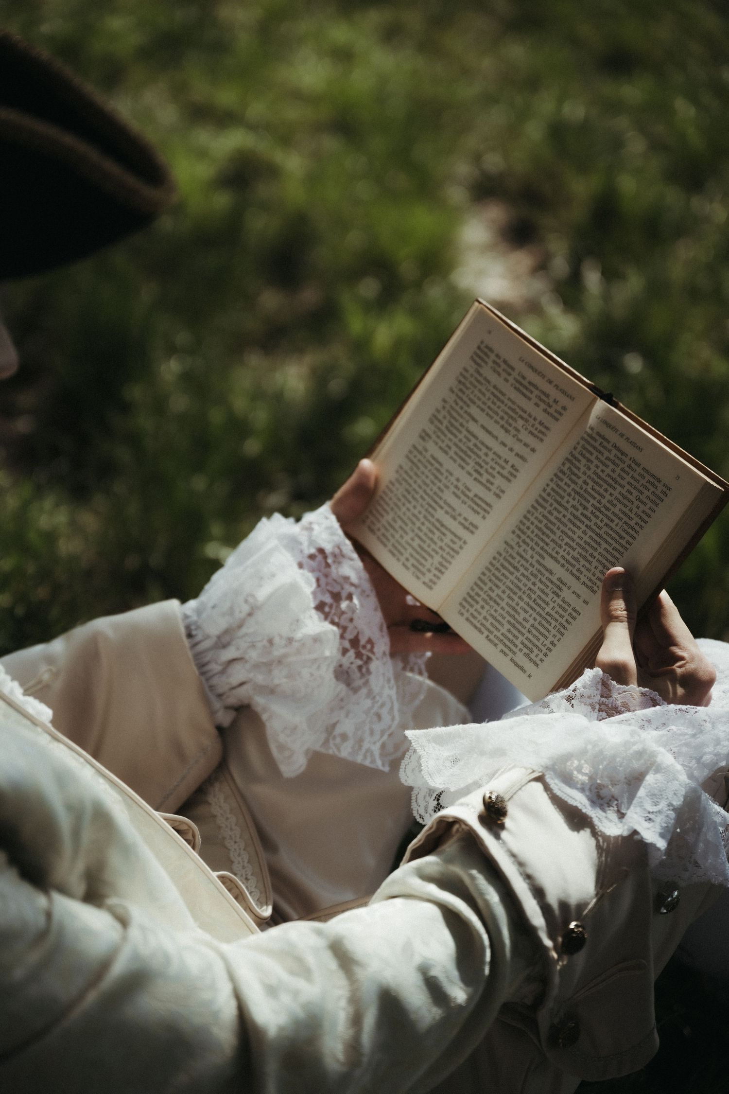 Person in vintage lace dress reading an open book outdoors on green grass in soft natural light.