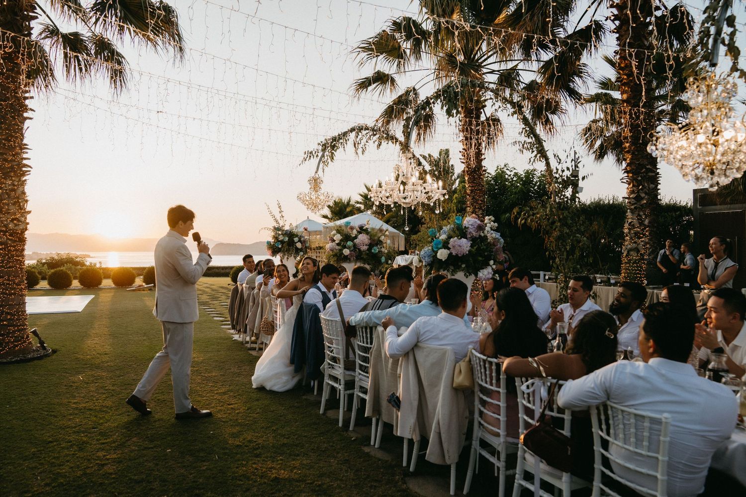 Guests seated at white chairs during an outdoor sunset wedding ceremony with palm trees in the background.