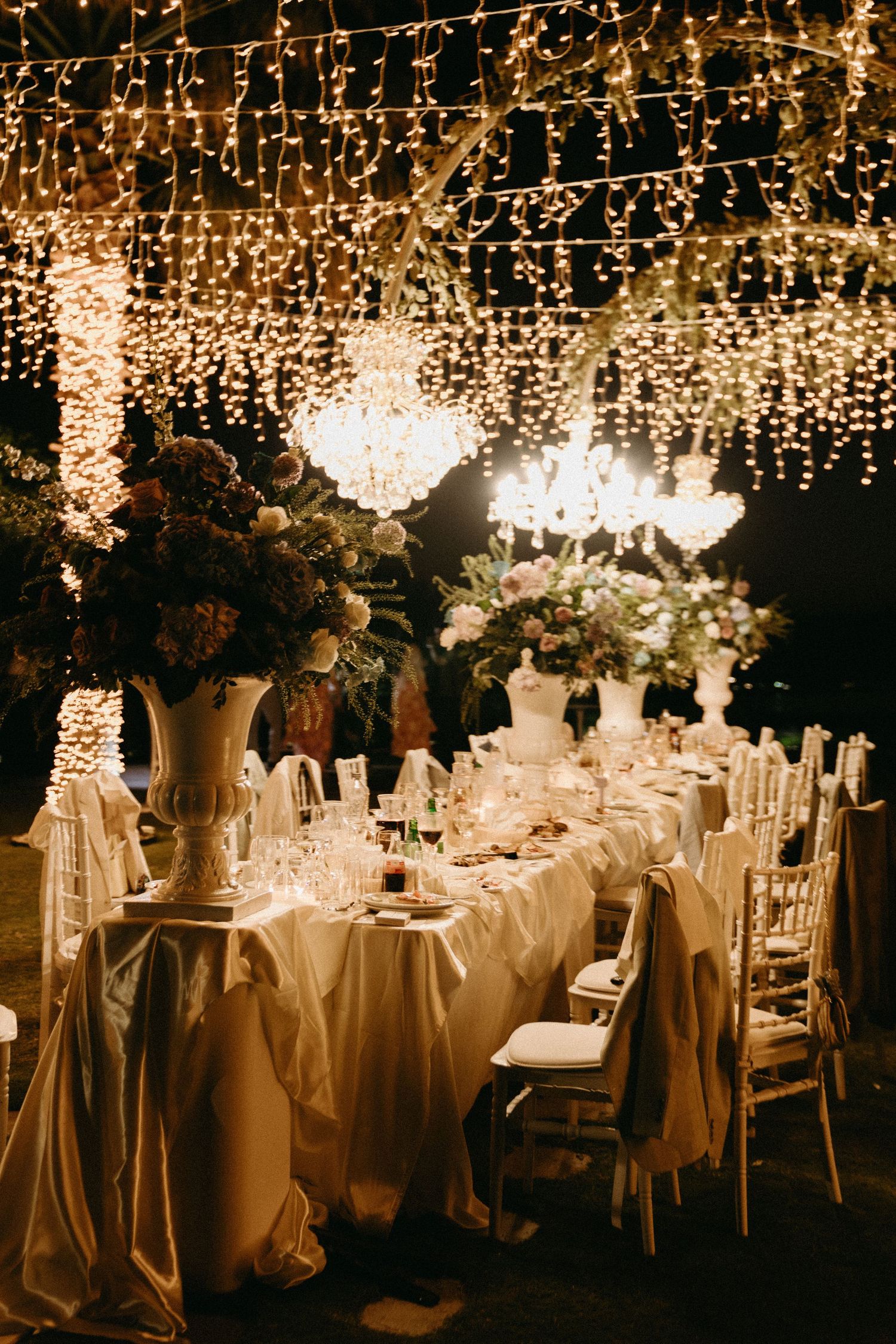 Elegant wedding reception table setting under a canopy of twinkling fairy lights with crystal chandeliers and floral centerpieces.