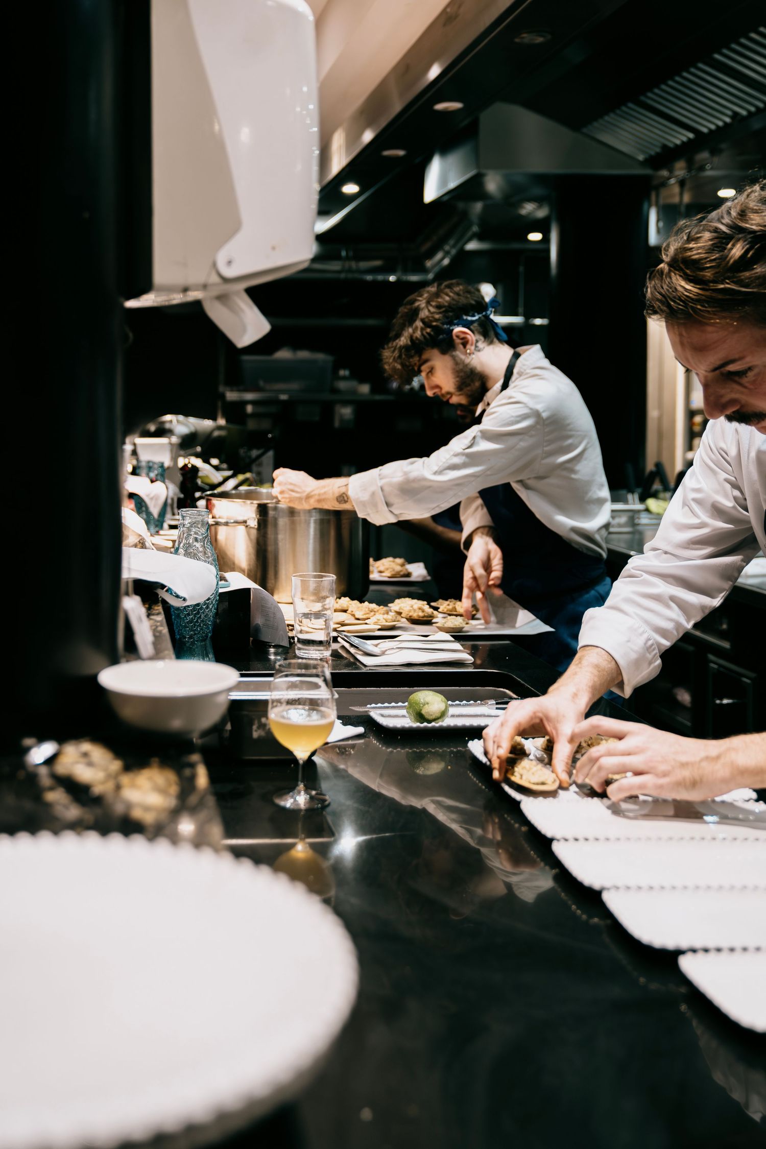 Chefs in white coats prepare dishes at a dark restaurant counter with focused precision and attention to detail.