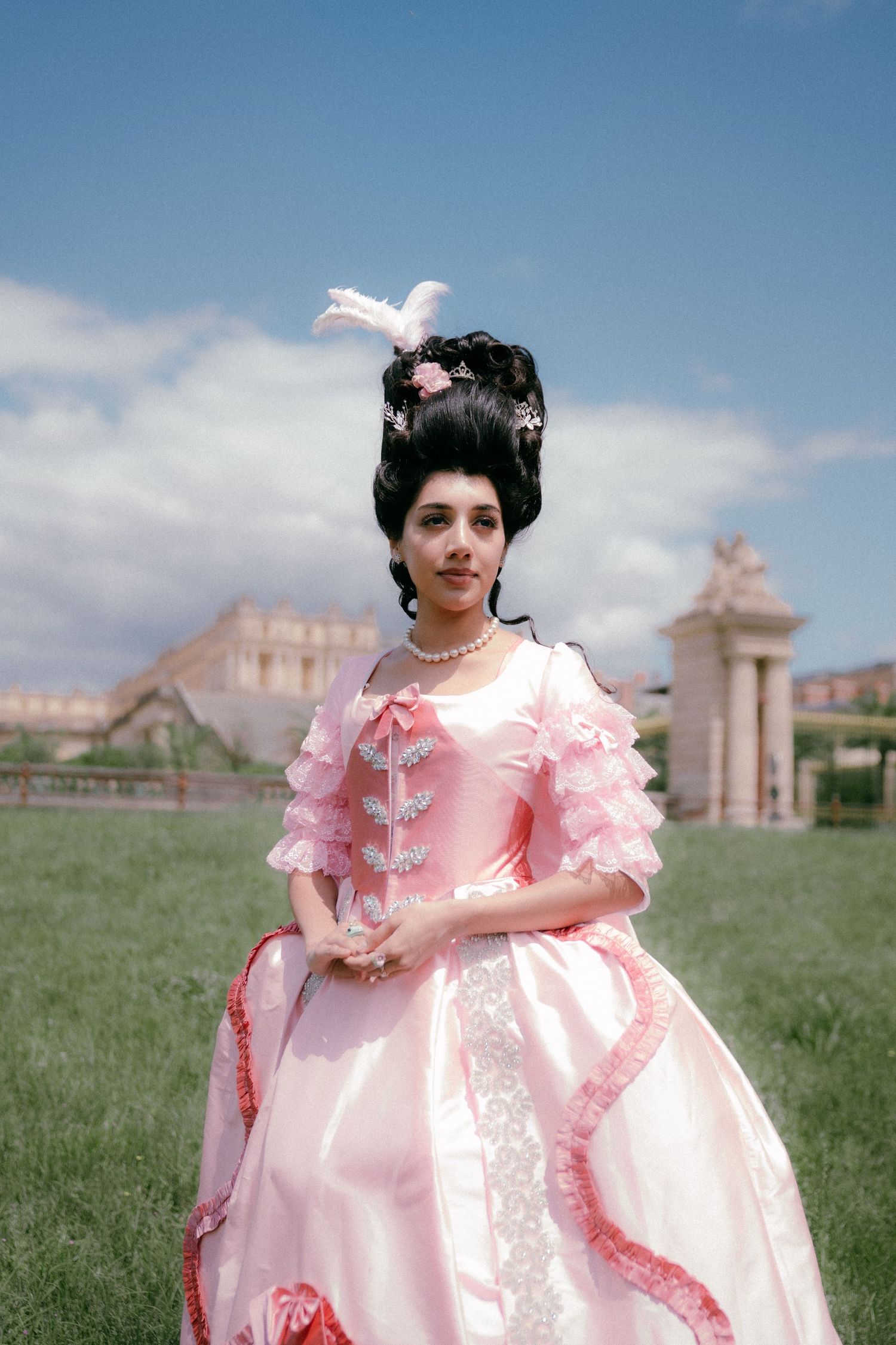 A person in a voluminous pink Victorian-style dress with ruffles poses in a grassy field with buildings in the background.