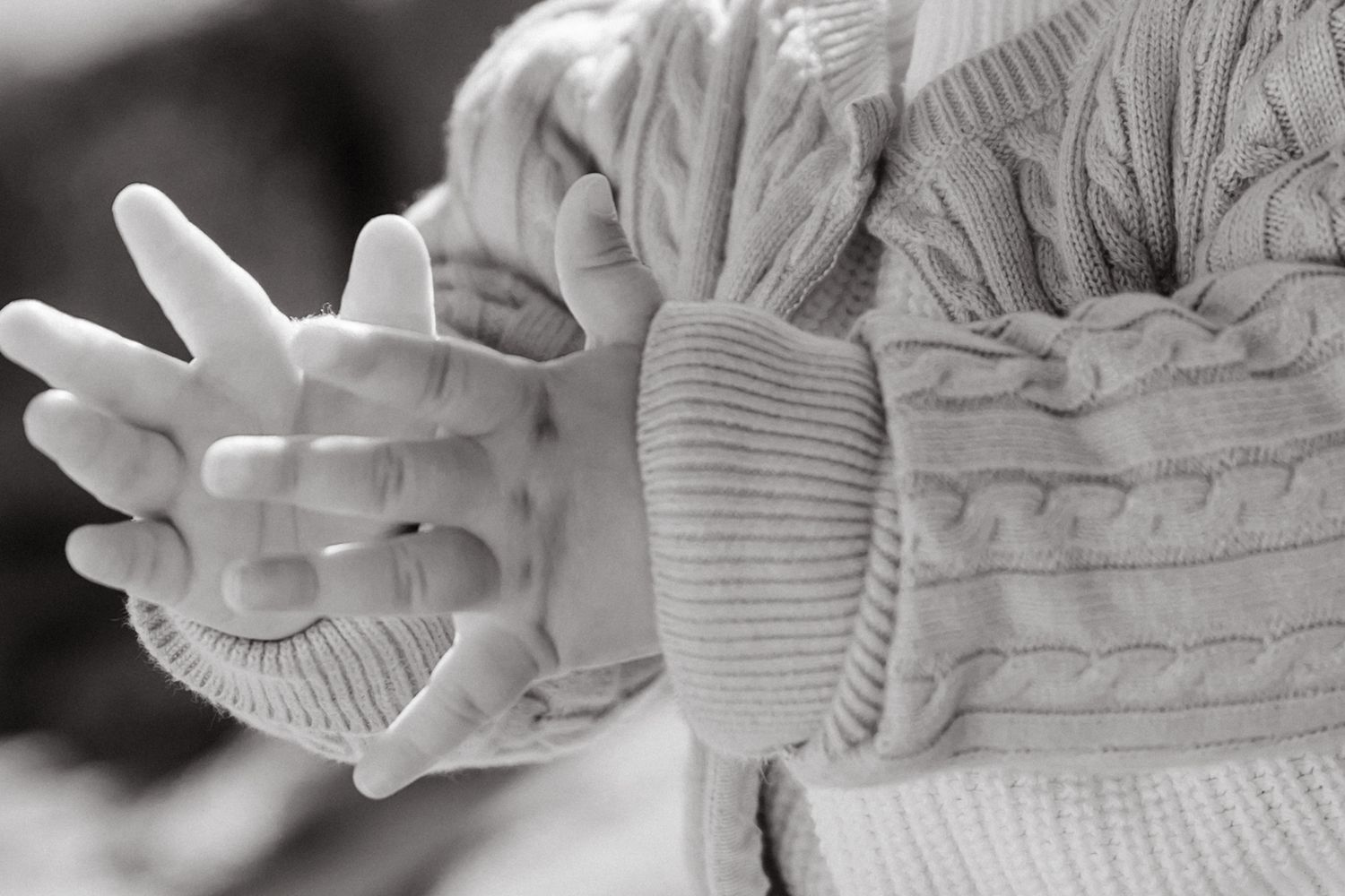 Black and white close-up of a baby's hands clasped together, wearing a cozy cable-knit sweater.