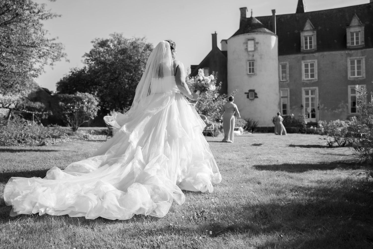 Bride walking across sunlit garden in front of a historic château near Paris, her voluminous gown trailing behind her as guests gather ahead