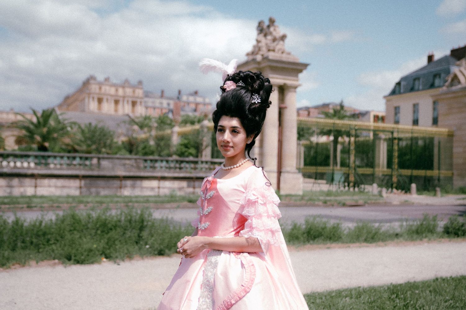 A person wearing a flowing pink historical gown stands in a formal garden with classical architecture in the background.