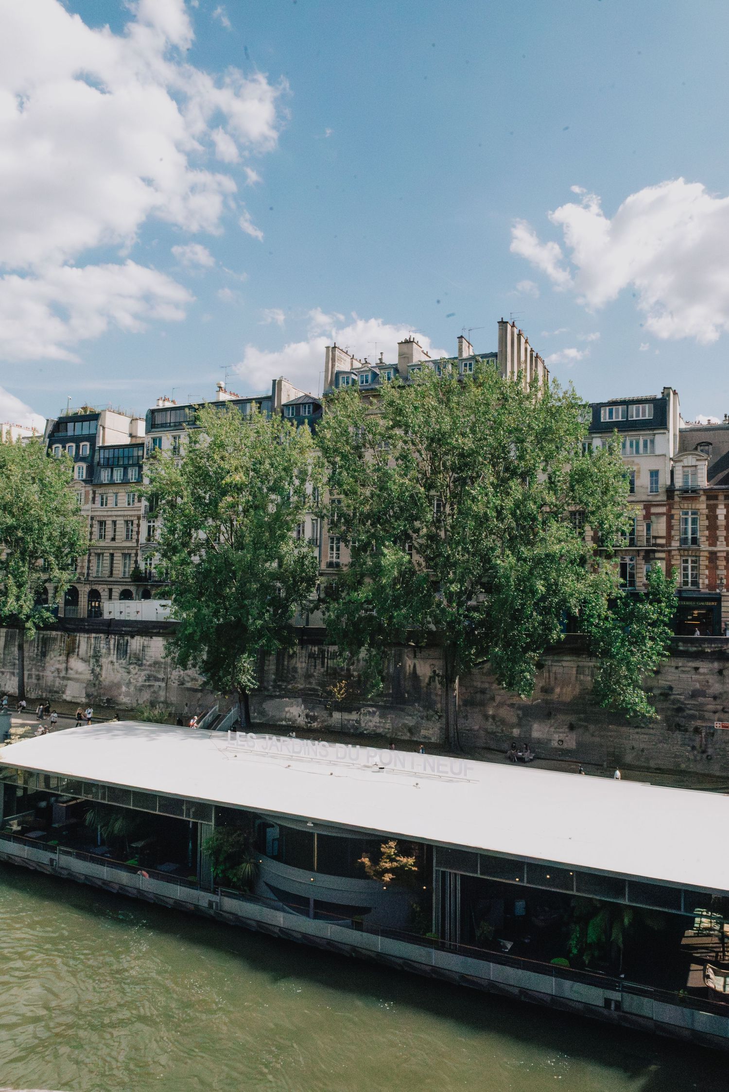 A panoramic view of historic buildings along a river embankment with trees and a white structure visible on a sunny day.