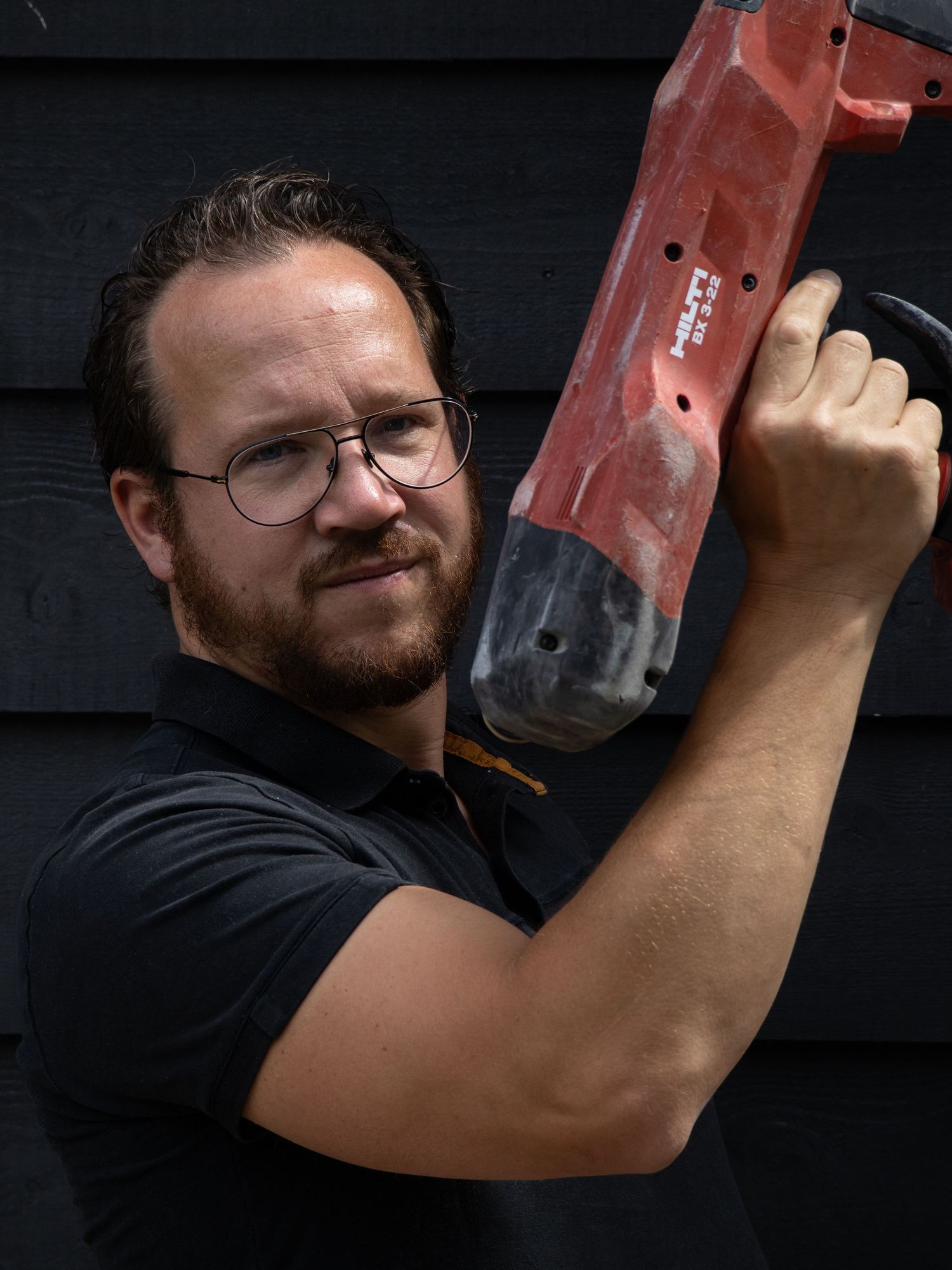 Man with glasses and beard holding a red Hilti power tool against a dark wooden wall outdoors.