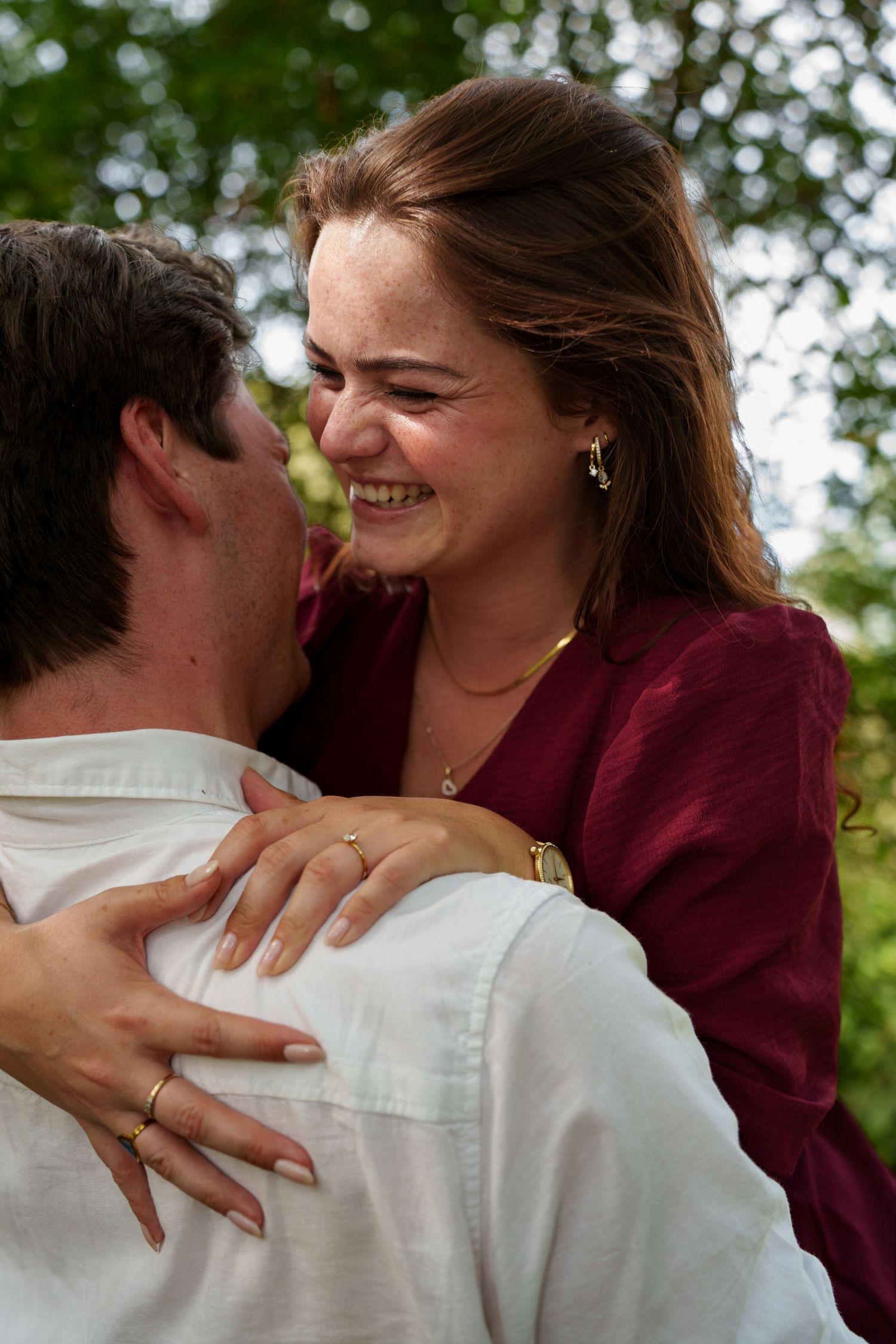 Happy couple embracing outdoors, woman in burgundy top smiling joyfully, wearing gold jewelry and ring.