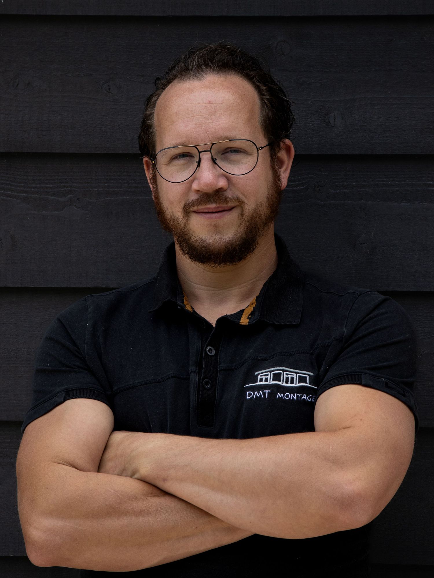 Man with glasses and crossed arms wearing a DMT Montage polo shirt, standing against a dark wooden wall.
