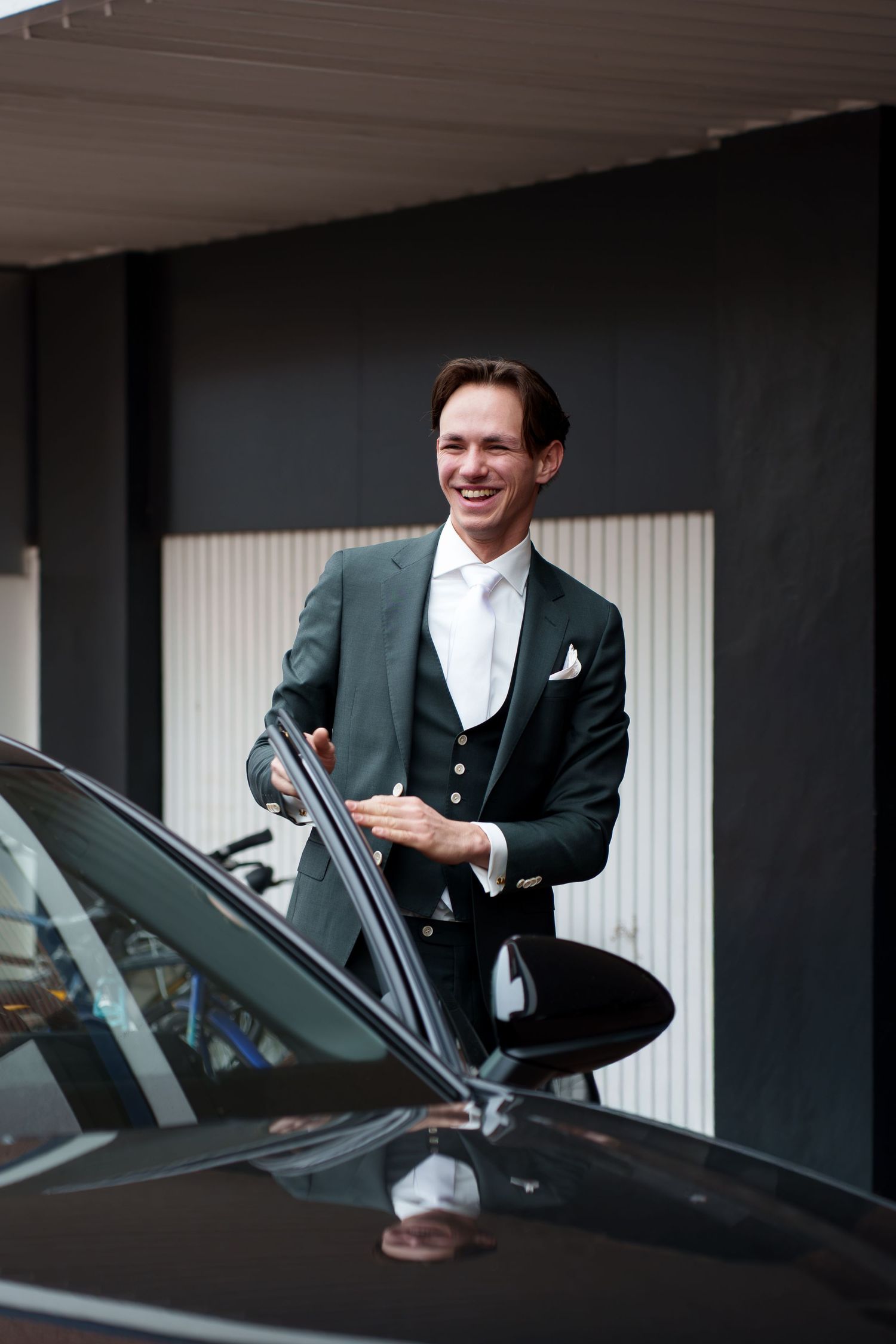 Smiling man in dark green suit and white tie opening a black car door outdoors.
