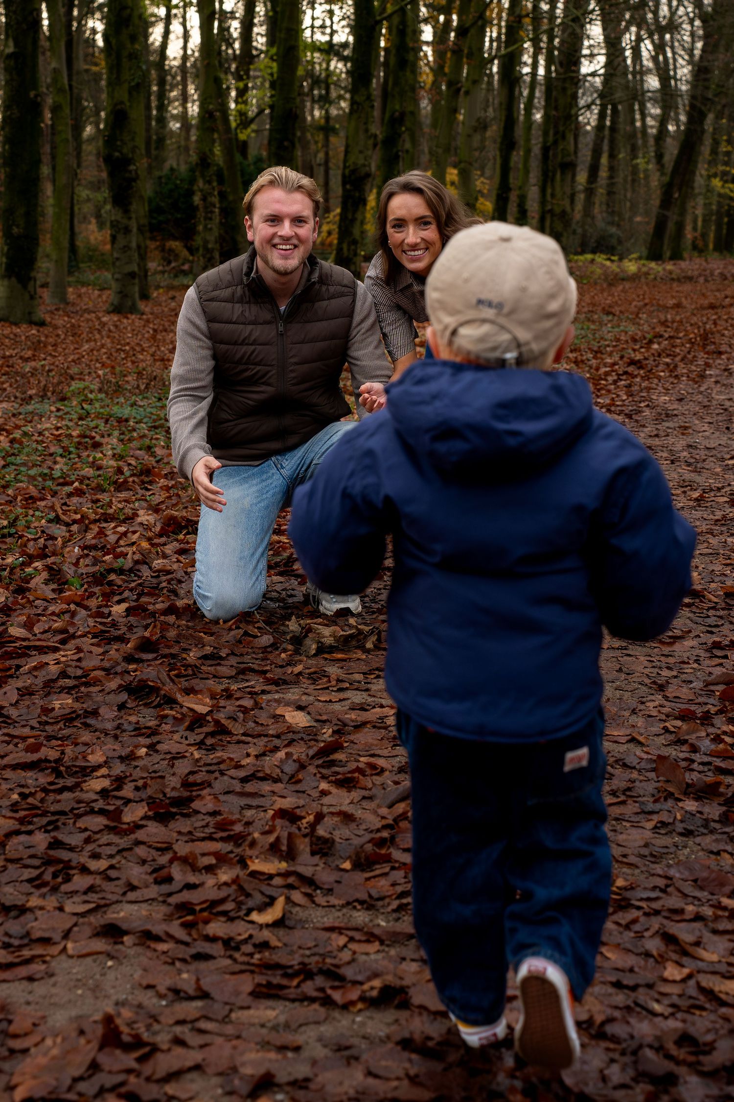 Young family playing in autumn forest, parents kneeling as toddler in blue jacket runs toward them through fallen leaves.