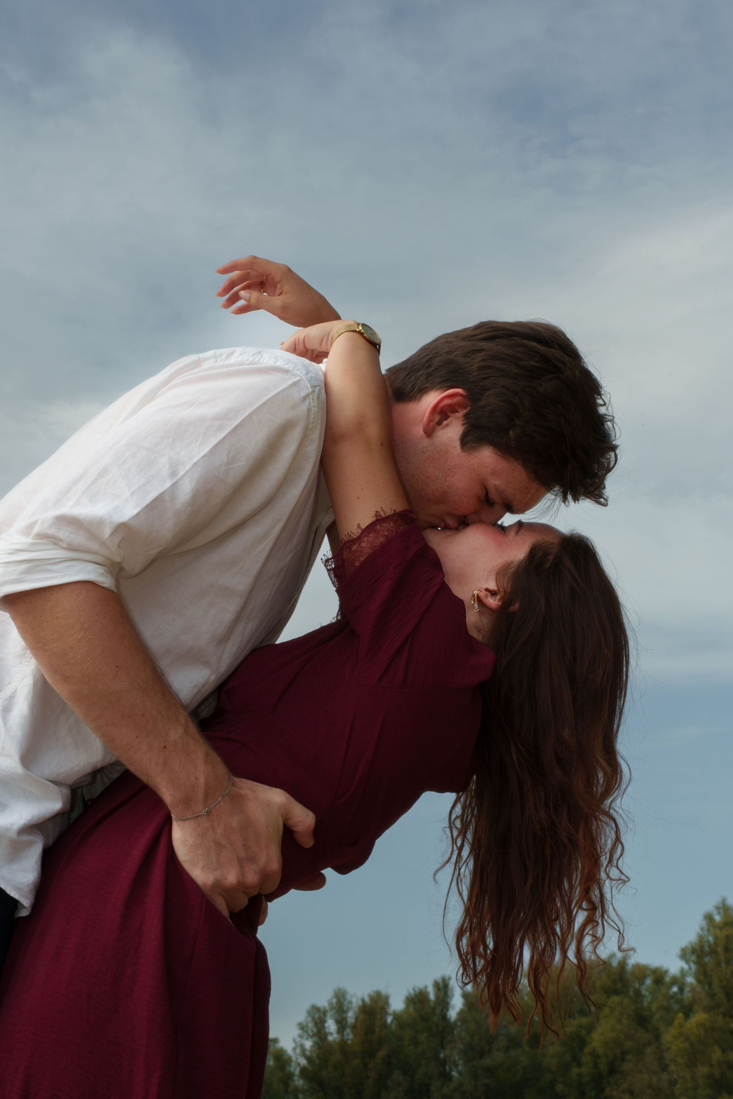 Romantic couple sharing a passionate kiss outdoors, man dipping woman in burgundy dress against cloudy sky.