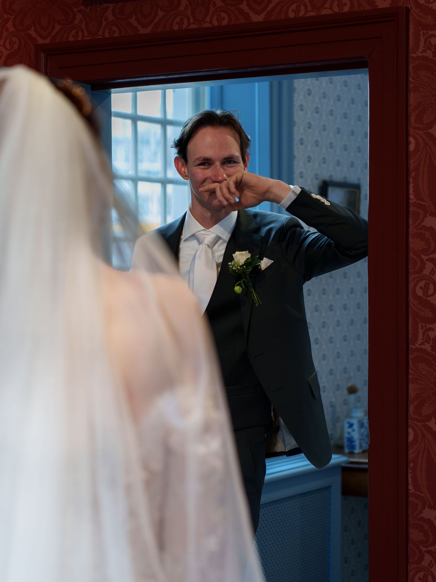 Emotional groom wiping tears seeing bride in wedding dress reflected in mirror during romantic first look moment.
