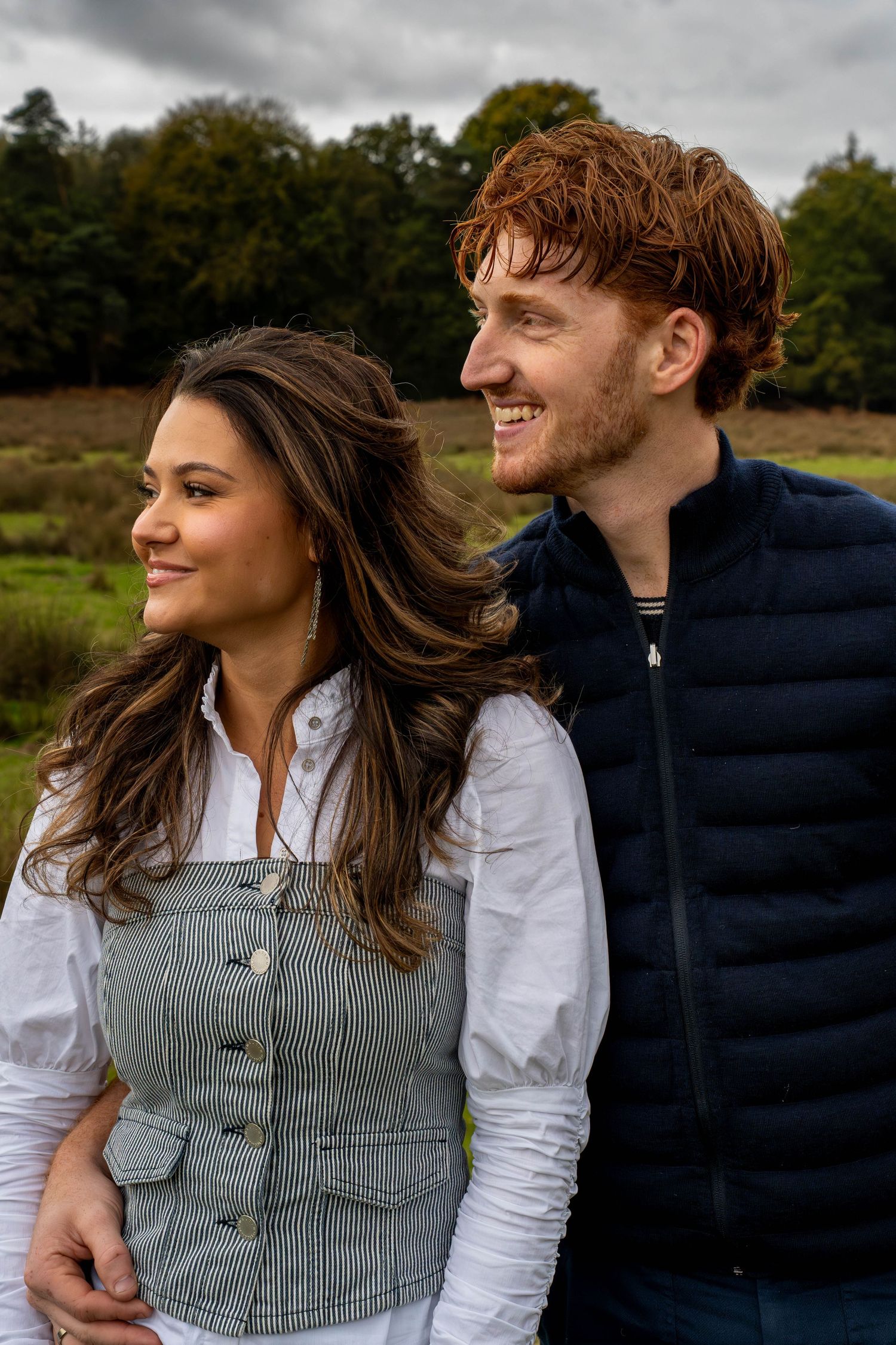 A couple in casual attire sits together outdoors, sharing a candid moment against a natural backdrop.