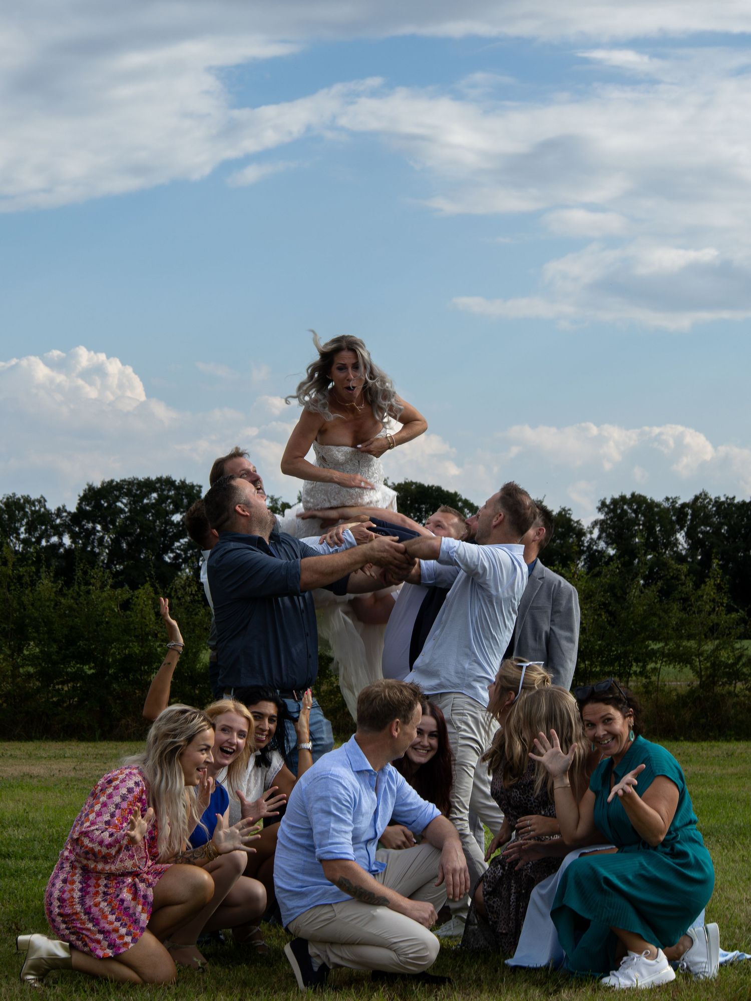Wedding guests joyfully tossing bride in the air outdoors on a sunny day with blue skies and green trees.