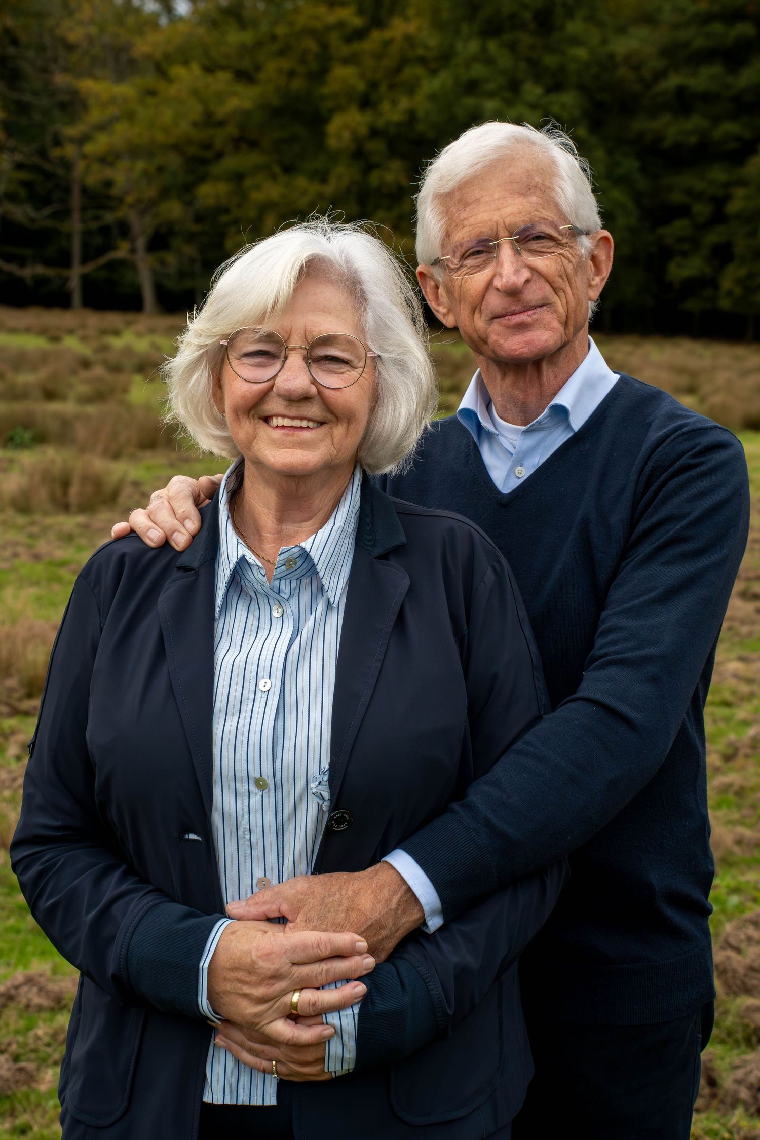 An elderly couple embraces outdoors on a sunny autumn day with trees in the background.
