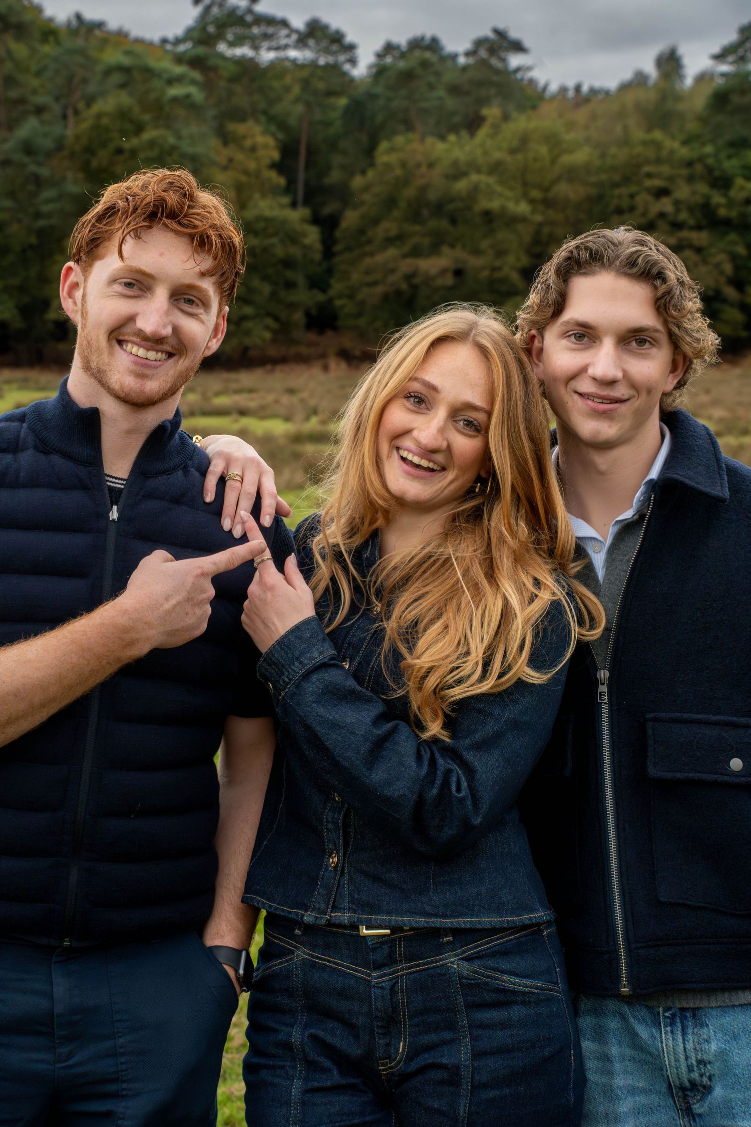 Three smiling young adults pose outdoors in denim outfits, with one man pointing at a ring on the woman's hand.