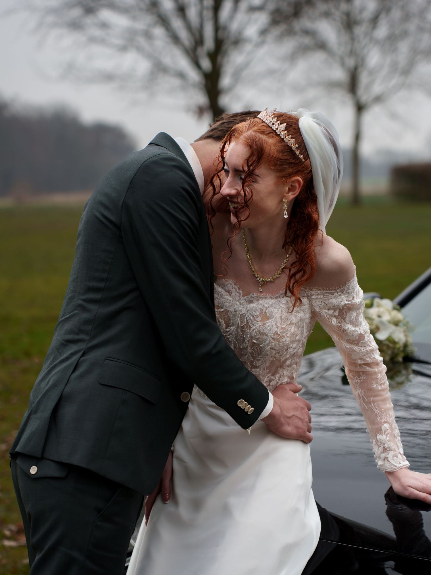 A bride in a lace off-shoulder gown and tiara shares a romantic moment with a groom in a dark suit by a car.