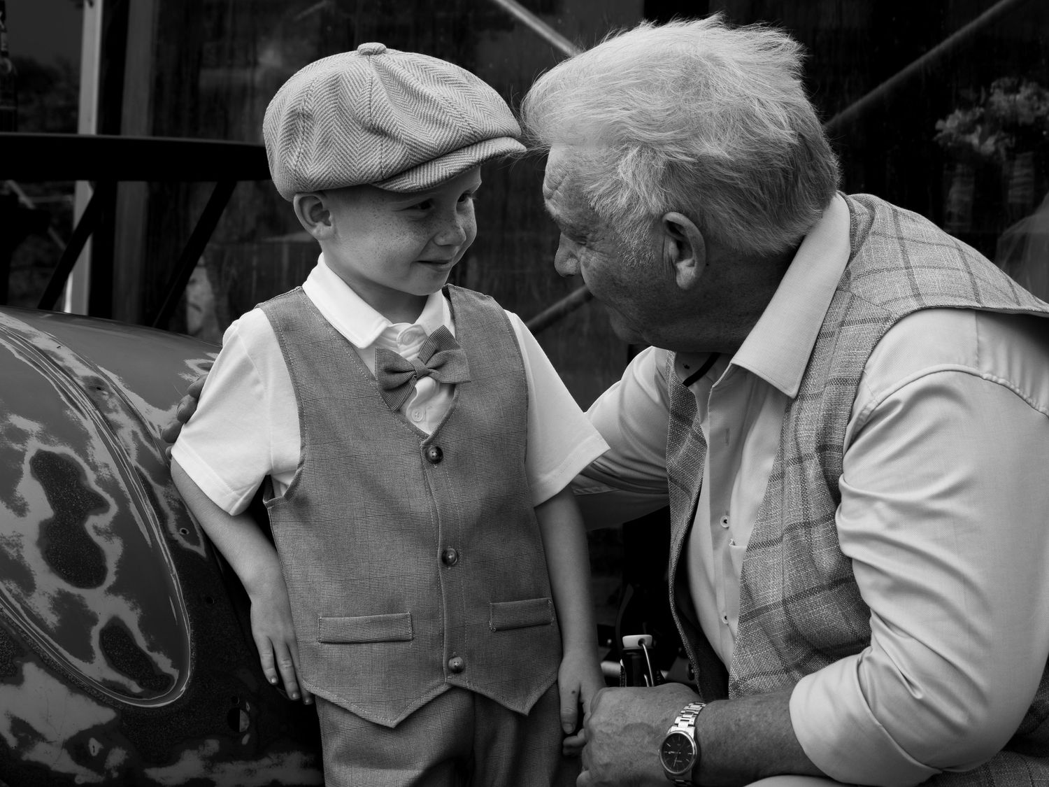 Black and white photo of a young boy in a vest and cap sharing a moment with an elderly man.