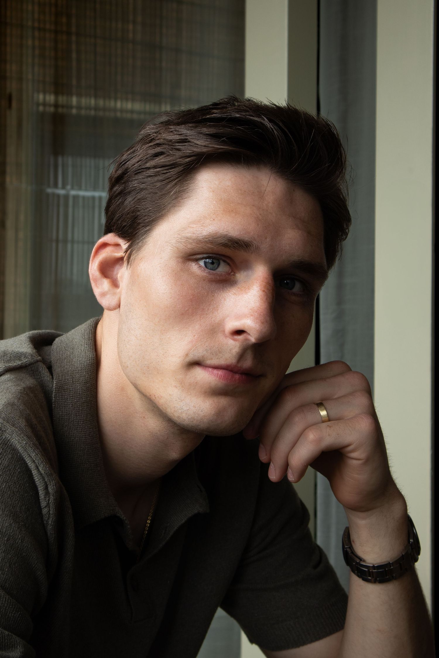 Portrait of person in casual hoodie near window with hand resting against chin, lit by natural light.