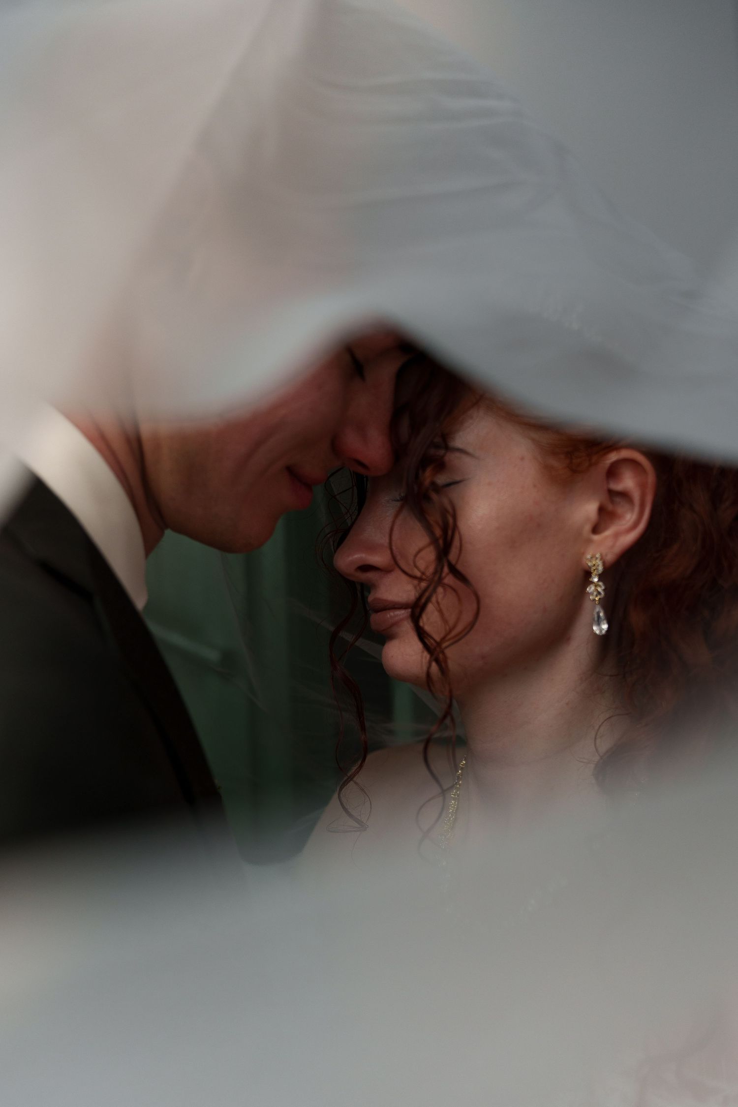 Bride and groom touch foreheads tenderly beneath a flowing white veil, sharing an intimate wedding moment.