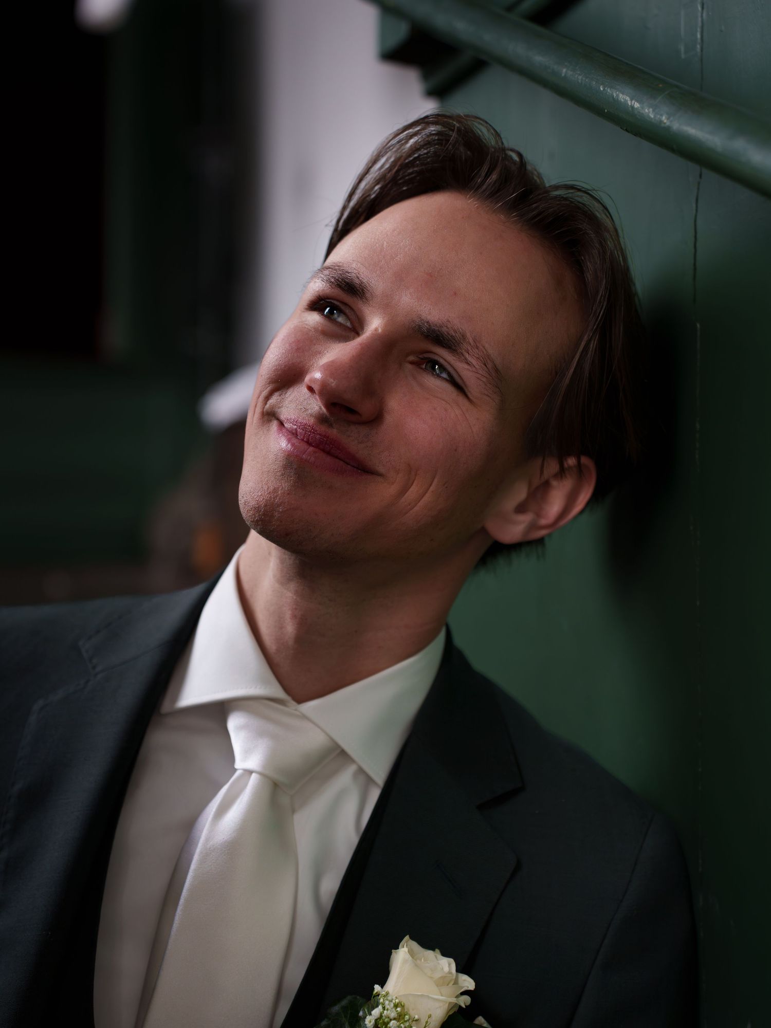 Young man in dark suit with white tie and boutonniere smiling upward at a formal wedding event.