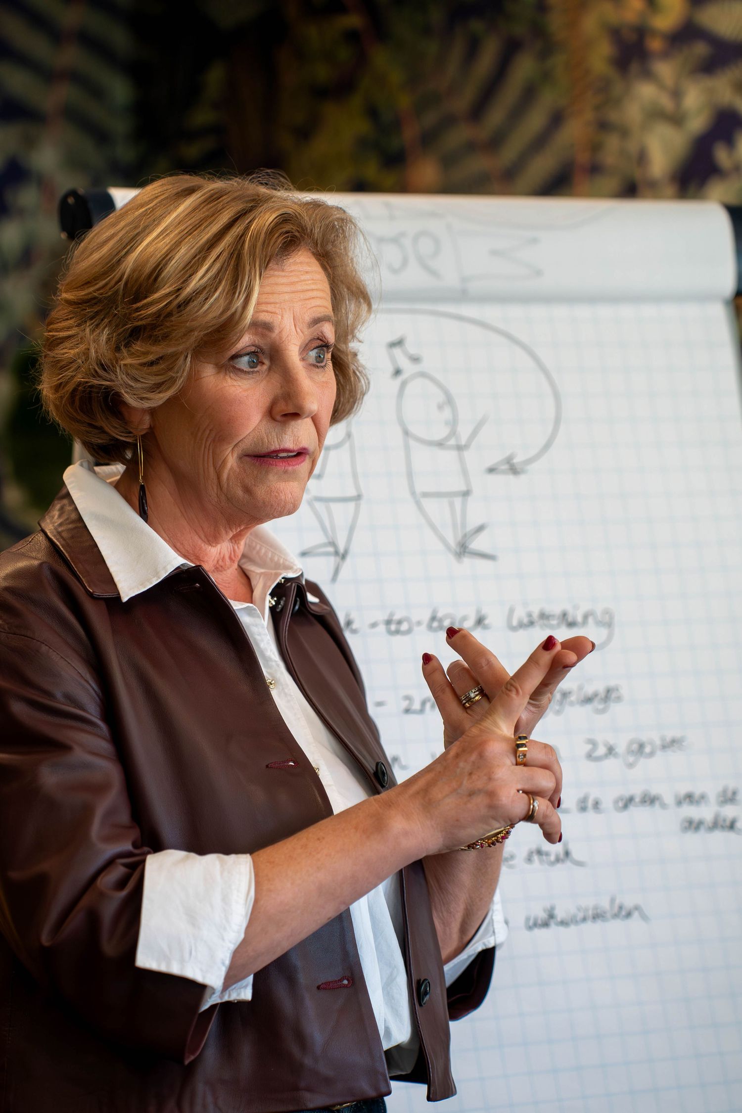 A woman in a brown blazer gestures while speaking in front of a whiteboard during a presentation.