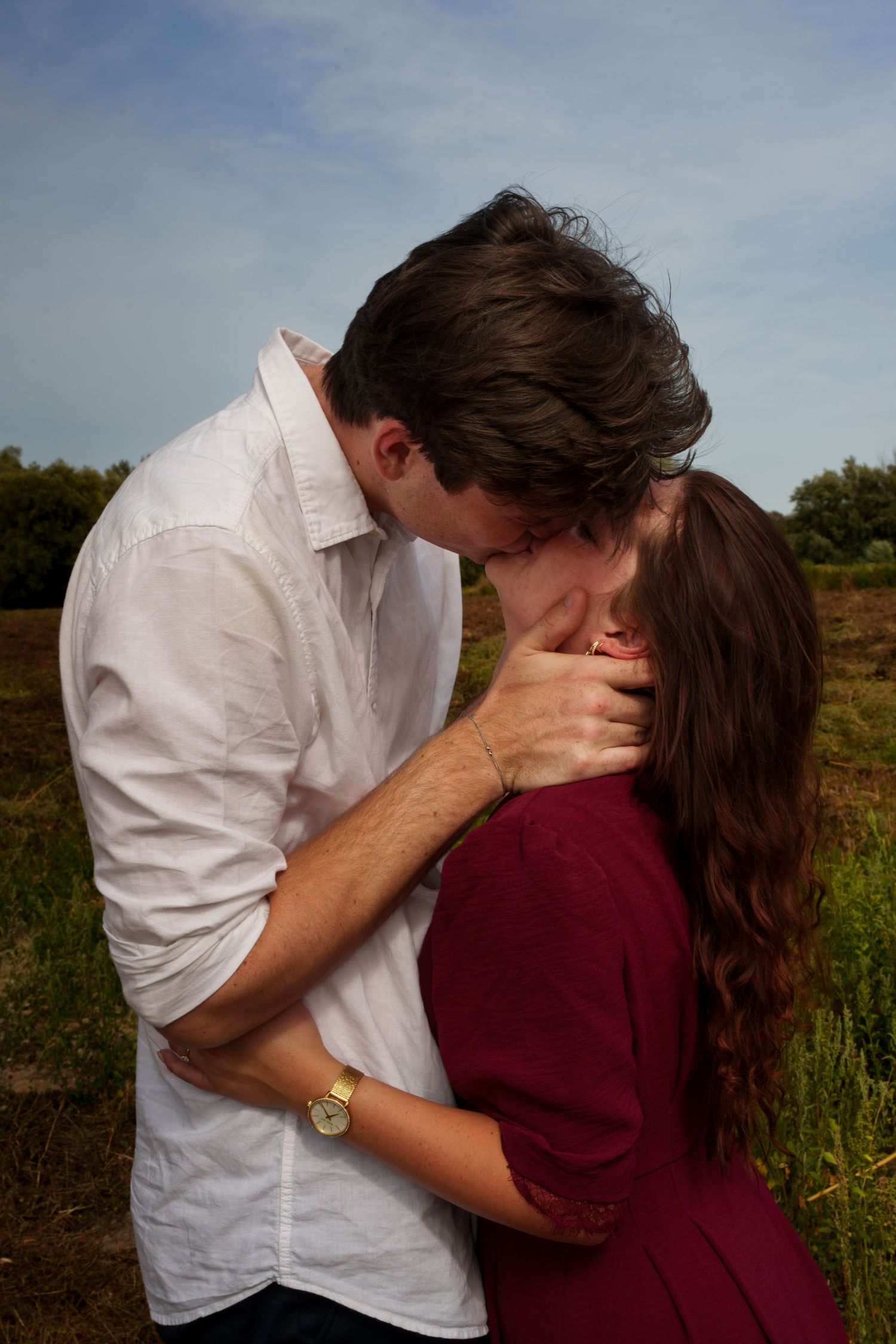 A couple sharing a romantic kiss in a green field, man in white shirt and woman in burgundy dress.