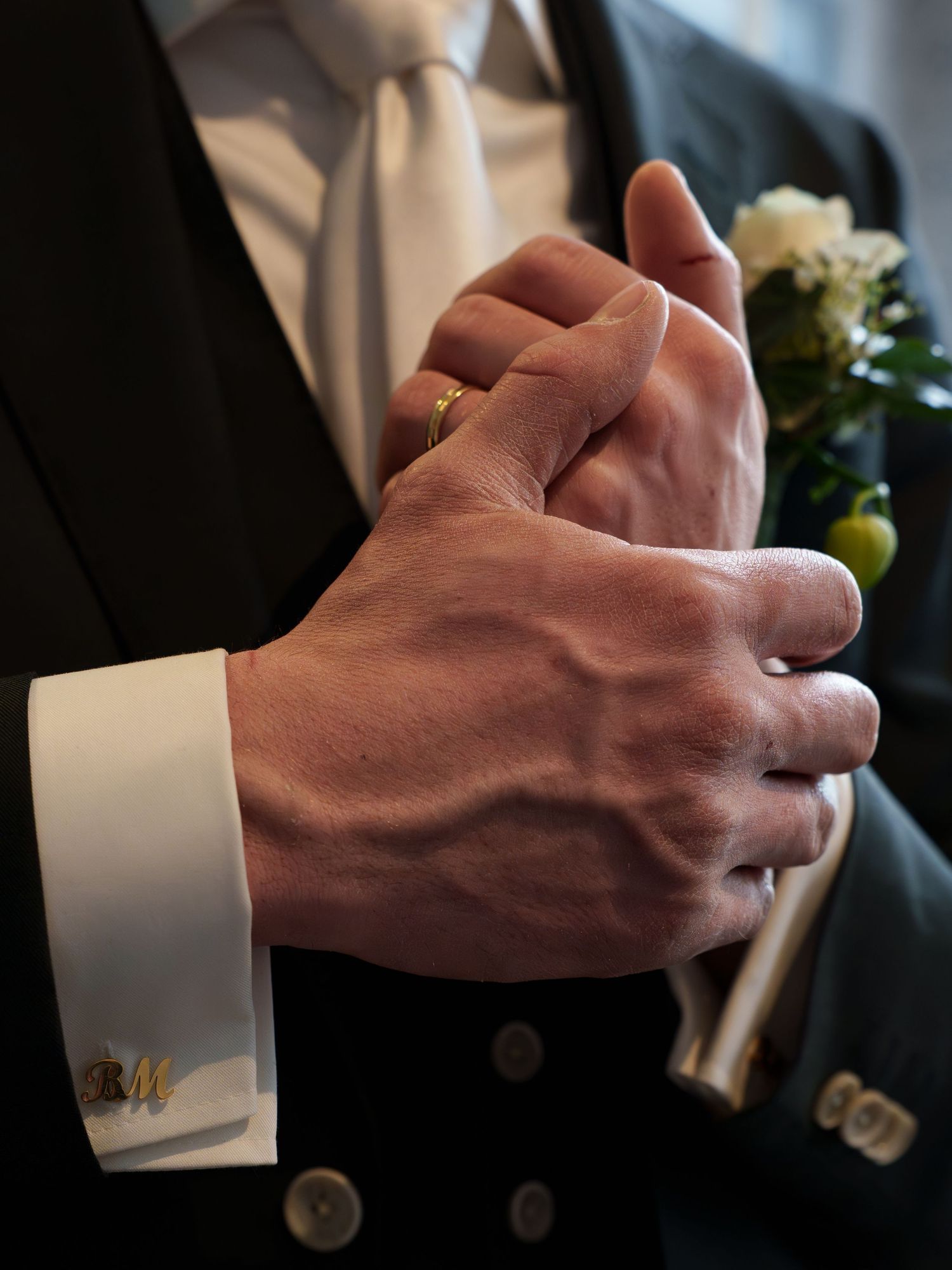 Close-up of a groom's hands with gold wedding ring, monogrammed cufflinks, white dress shirt, and floral boutonniere.
