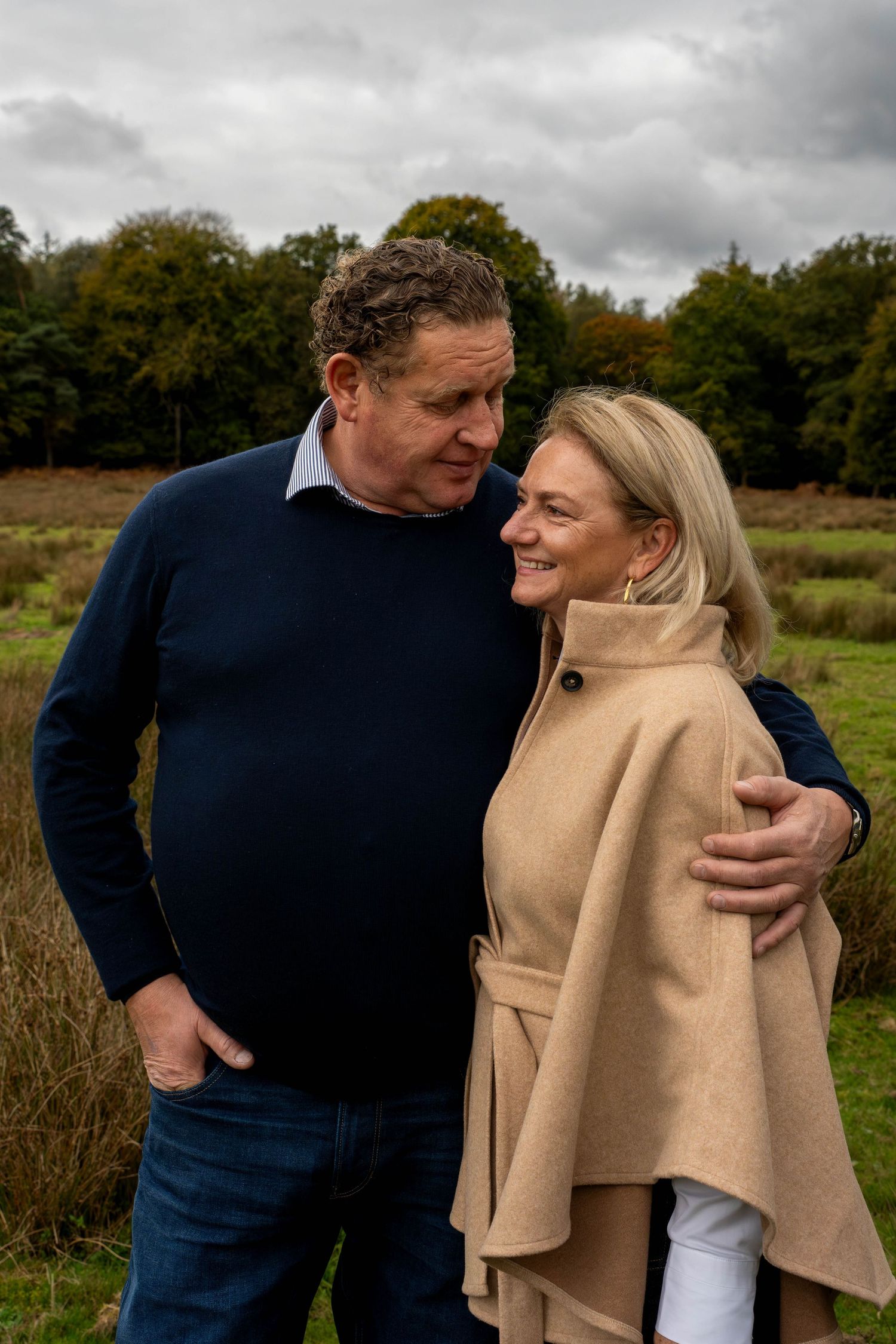 A middle-aged couple embracing outdoors in a field, smiling warmly, wearing casual autumn clothing on a cloudy day.