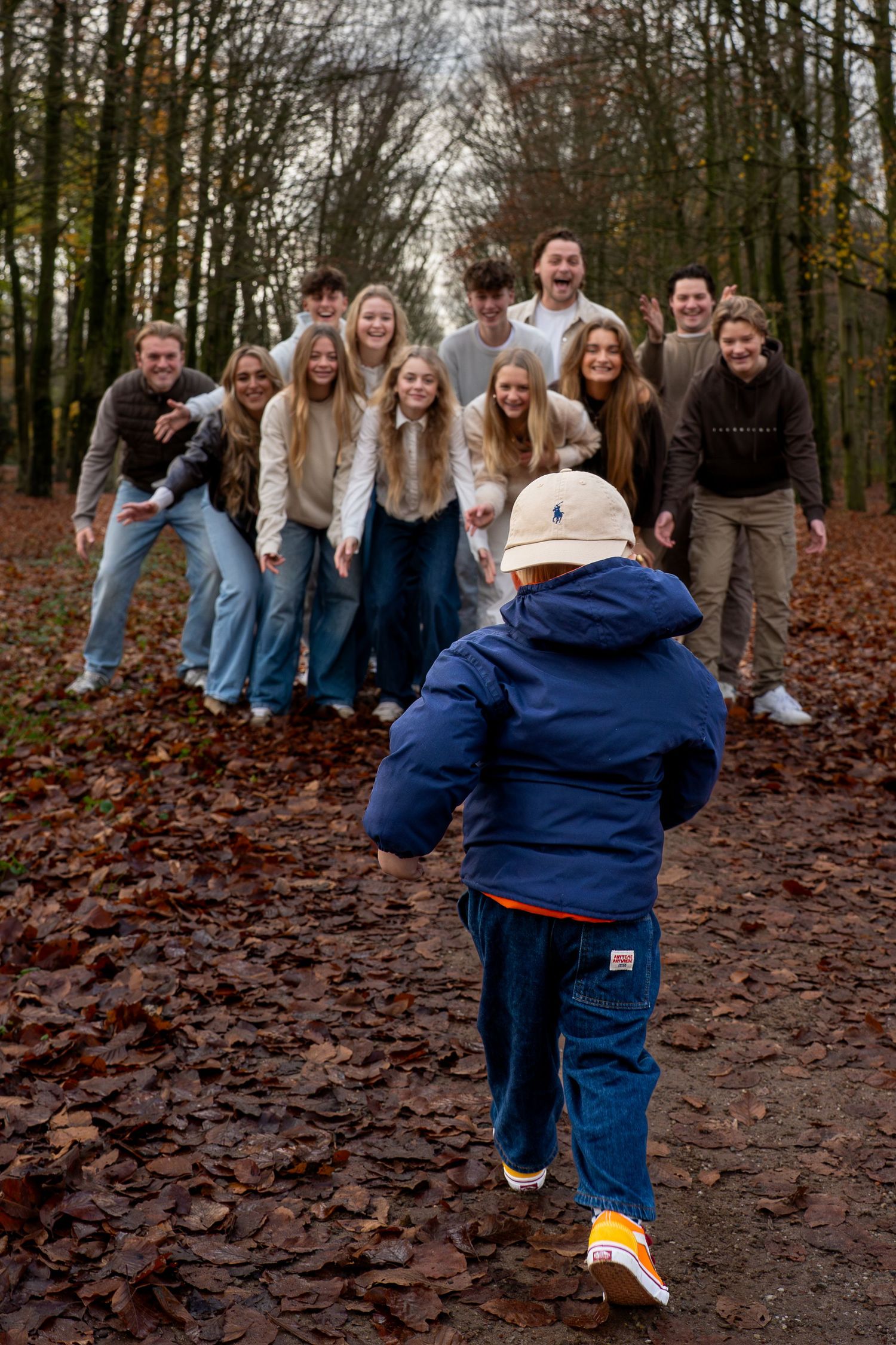 A young child in a blue jacket runs toward a group of people posing on a leaf-covered forest path in autumn.