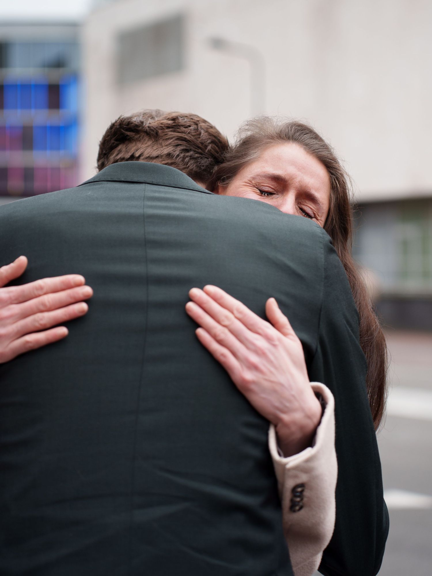 Two people sharing an emotional embrace outdoors, woman with long brown hair hugging a man in a dark suit.