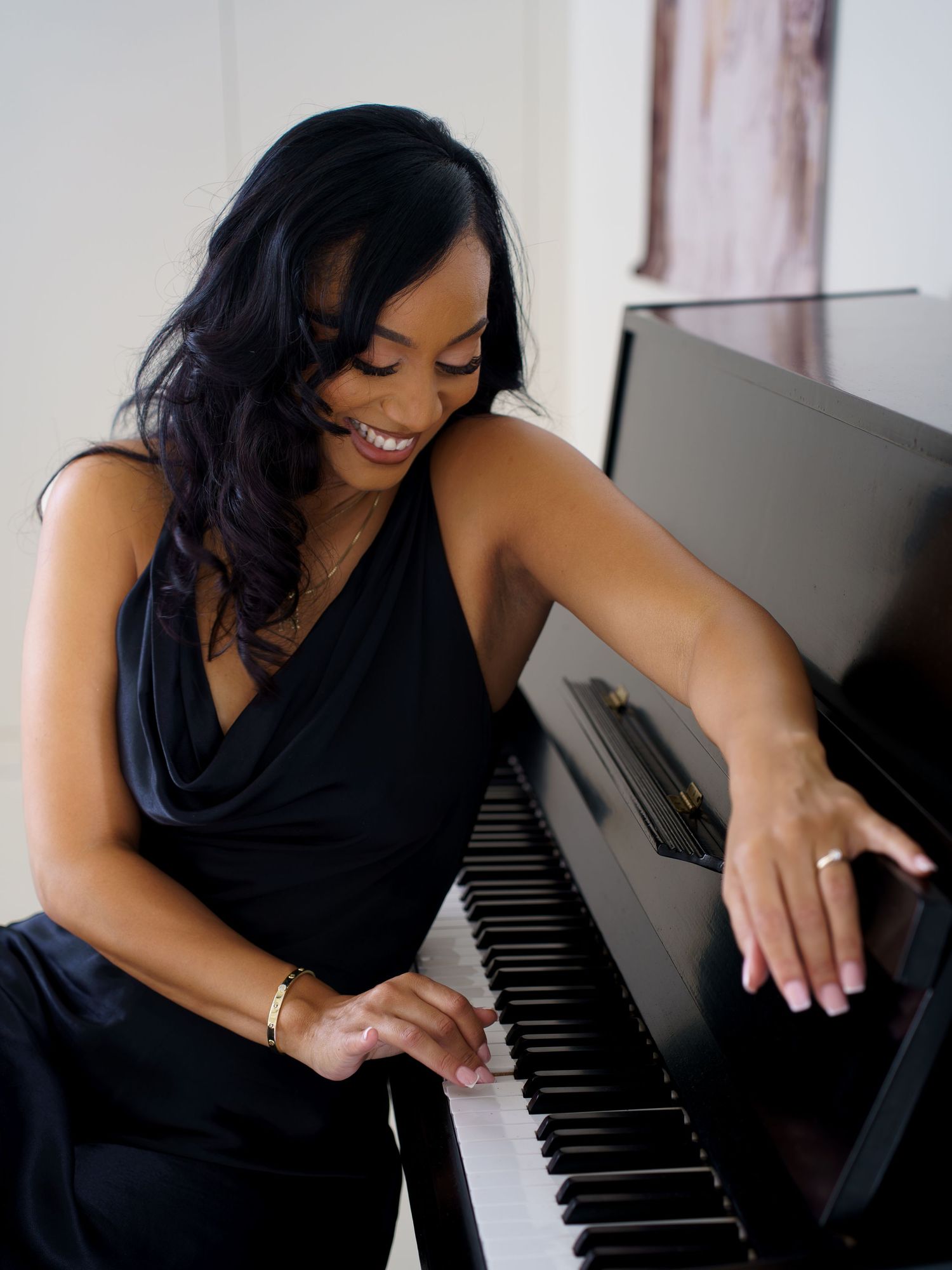 Smiling woman in black dress playing piano, showcasing elegance and musical talent in a bright, modern setting.