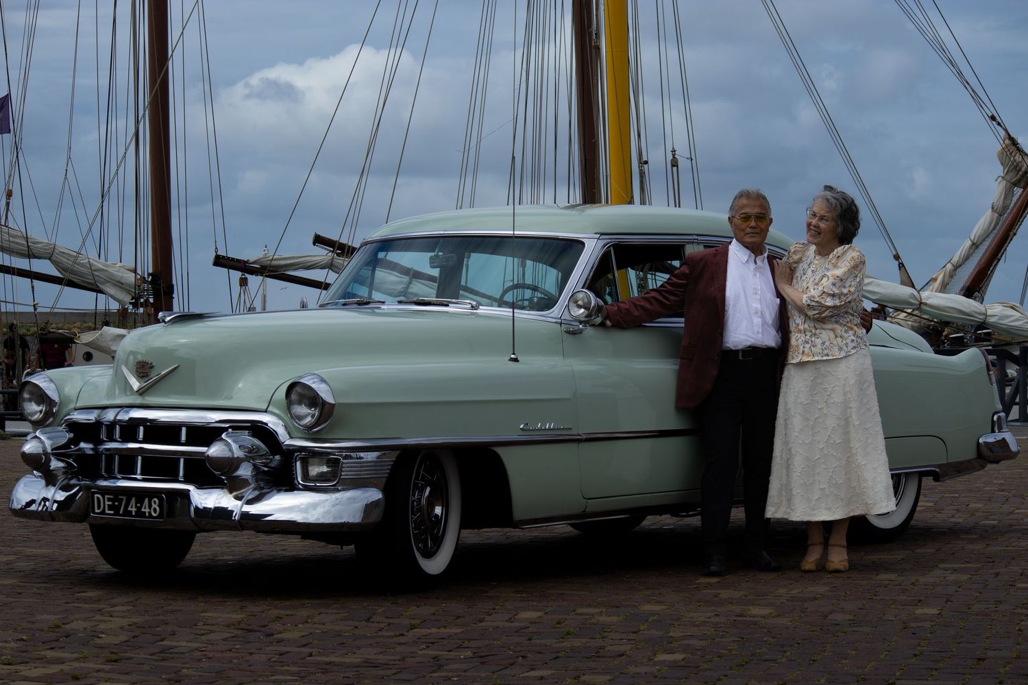 Elderly couple posing beside a classic mint green Cadillac on a cobblestone dock with tall ships in the background.