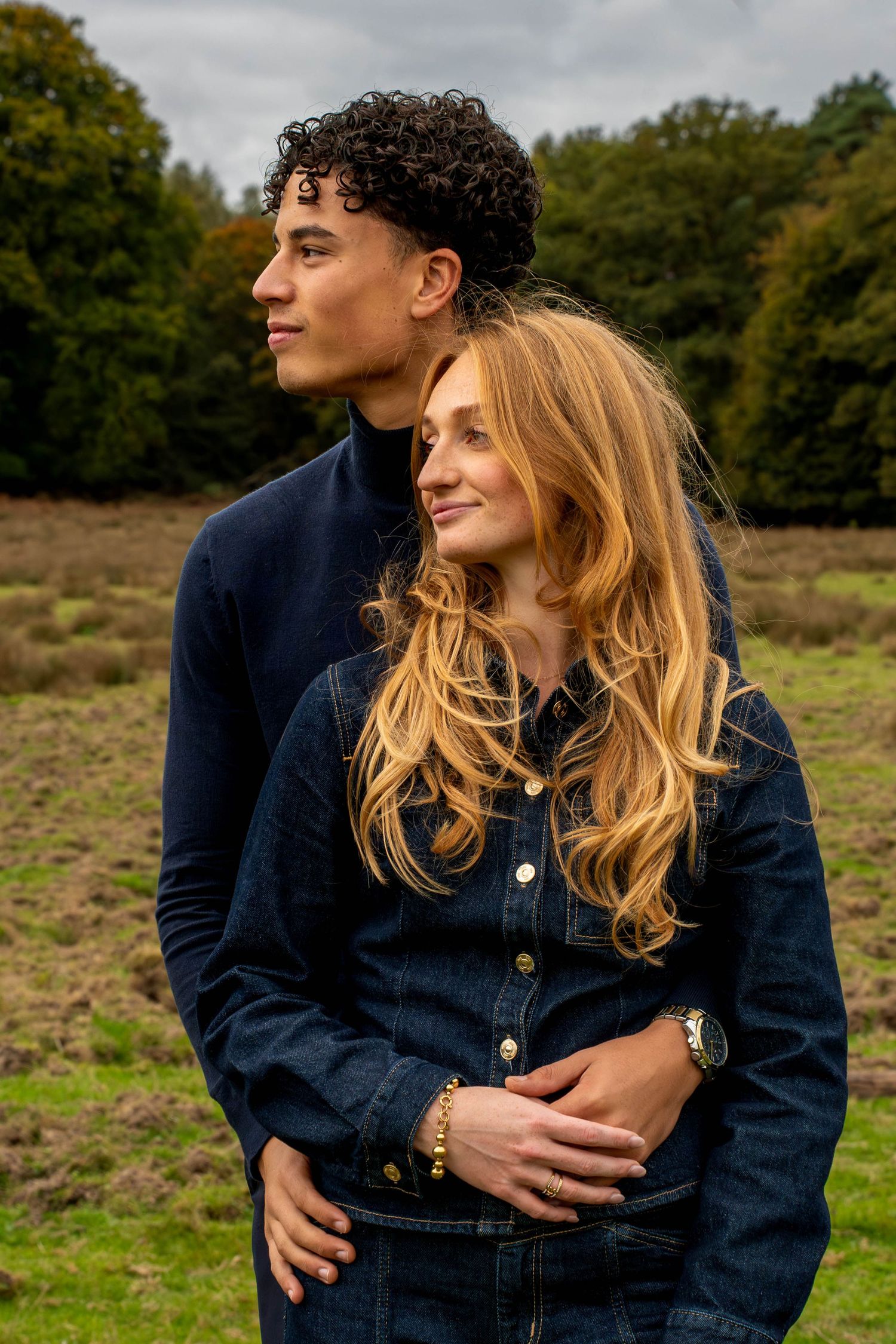 Young couple embracing outdoors in autumn, woman in denim jacket smiling, man in navy turtleneck, green trees behind.