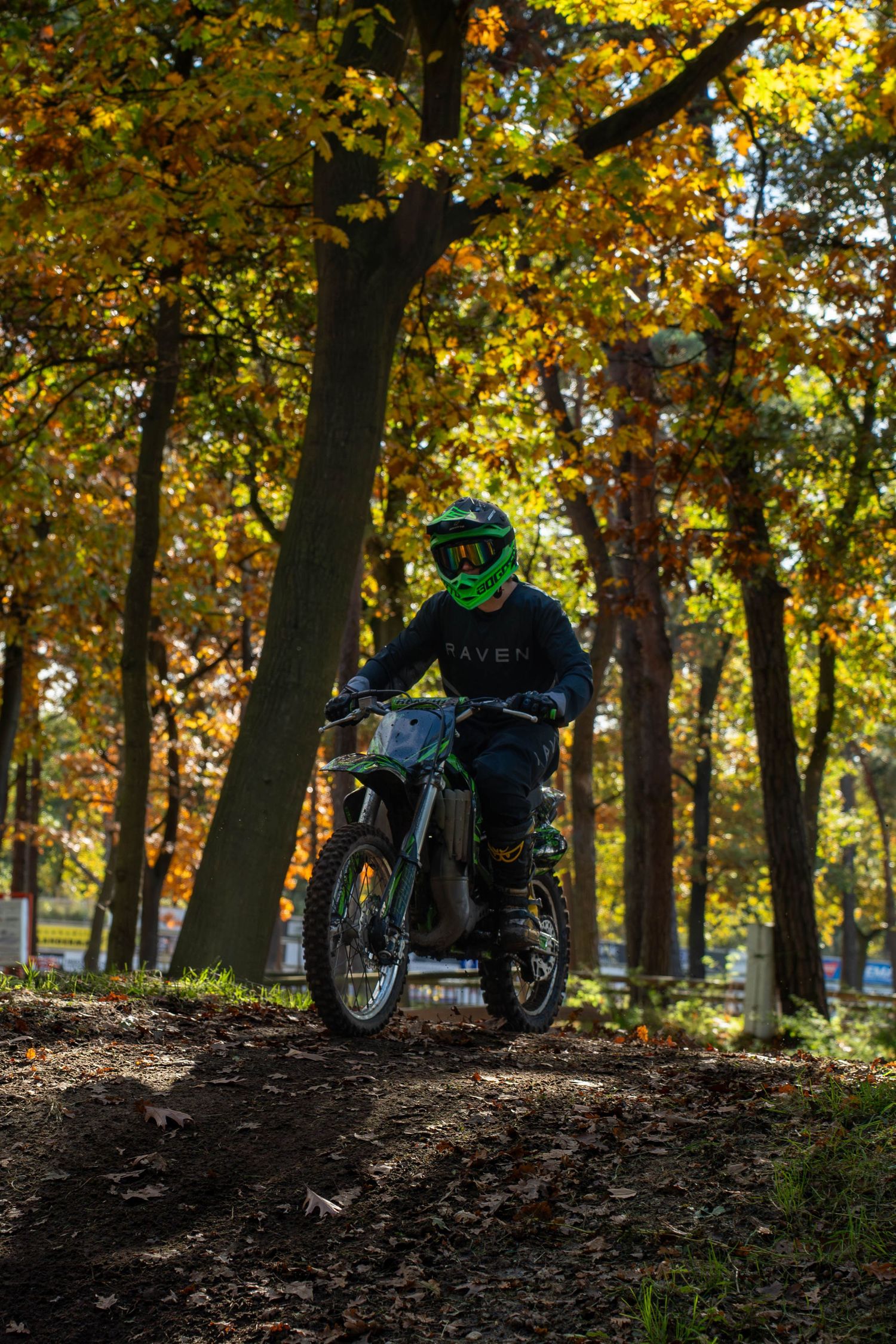 Motocross rider in green helmet navigates dirt trail through vibrant autumn forest with colorful fall foliage.