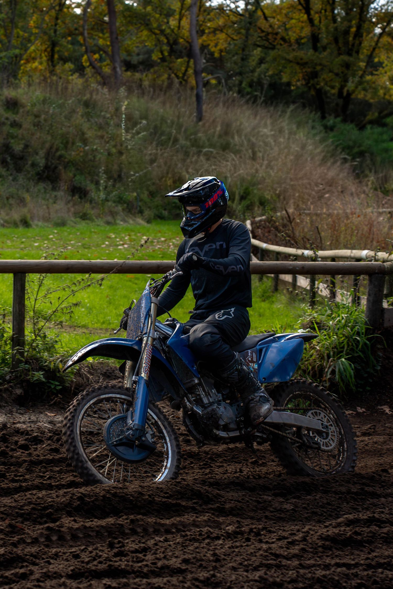 A dirt bike rider navigates through muddy terrain next to a wooden fence in a wooded area during an off-road session.