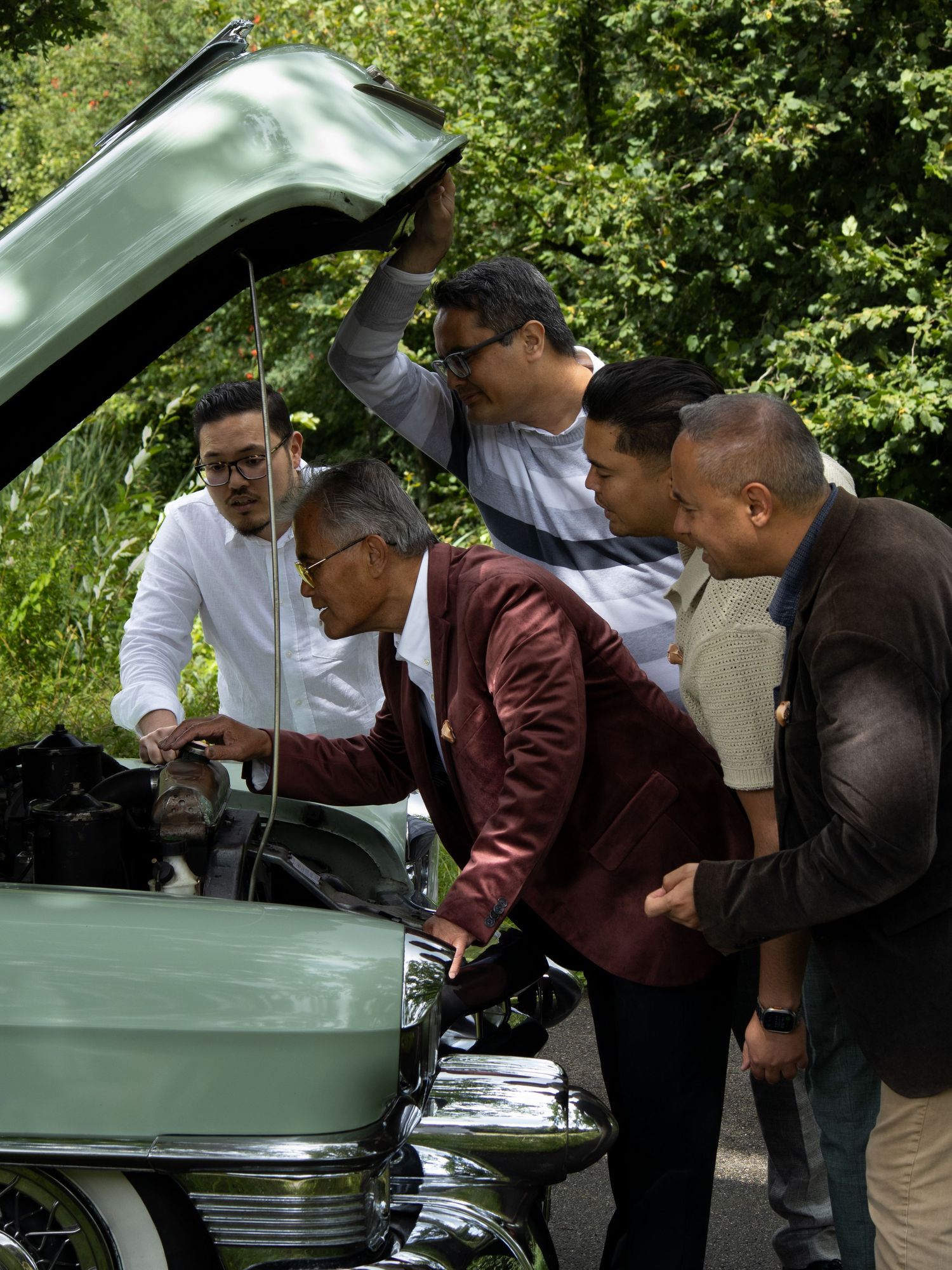 A group of people examining under the hood of a light green classic car outdoors.