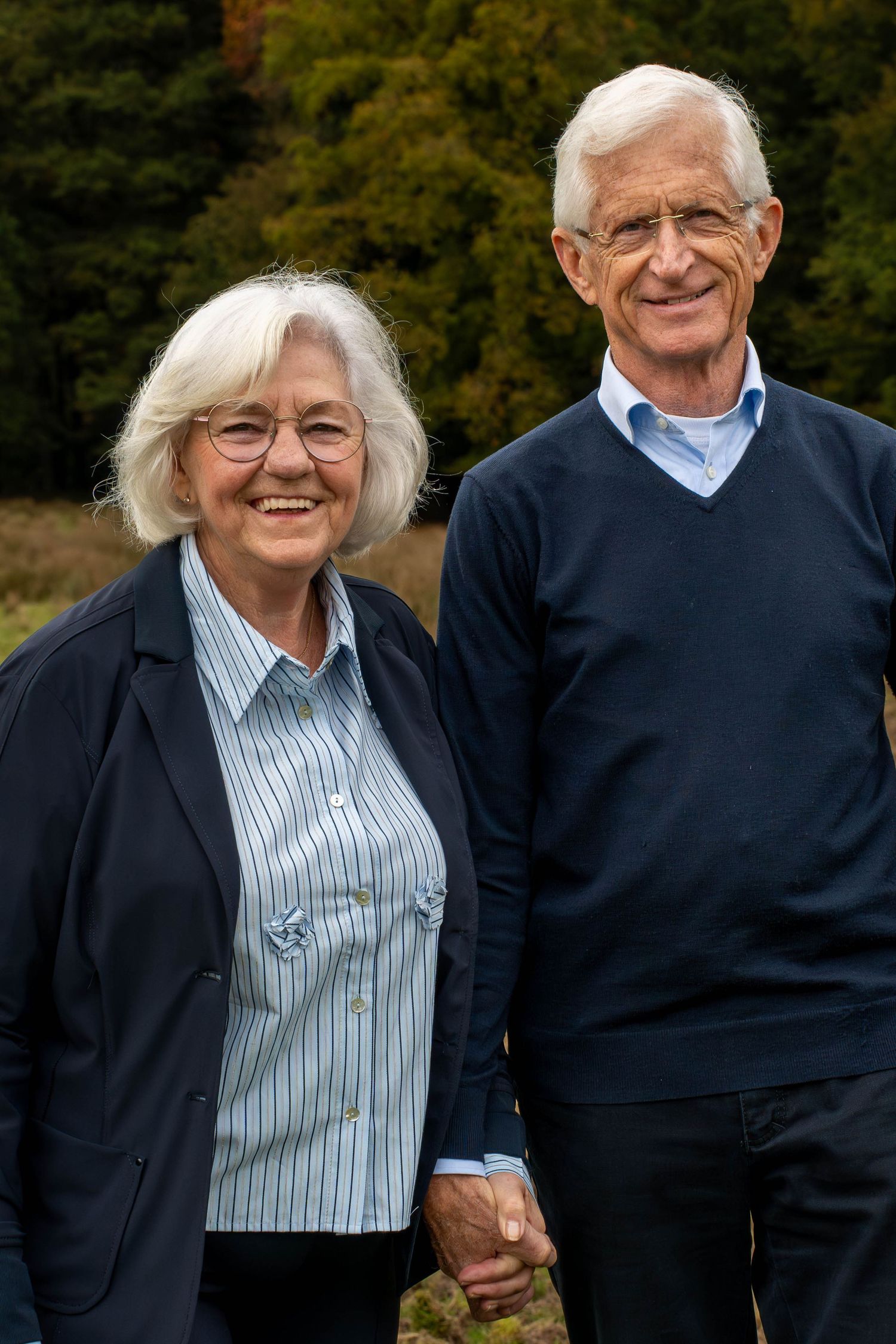 Smiling senior couple holding hands outdoors with green trees in background, wearing casual blue clothing.