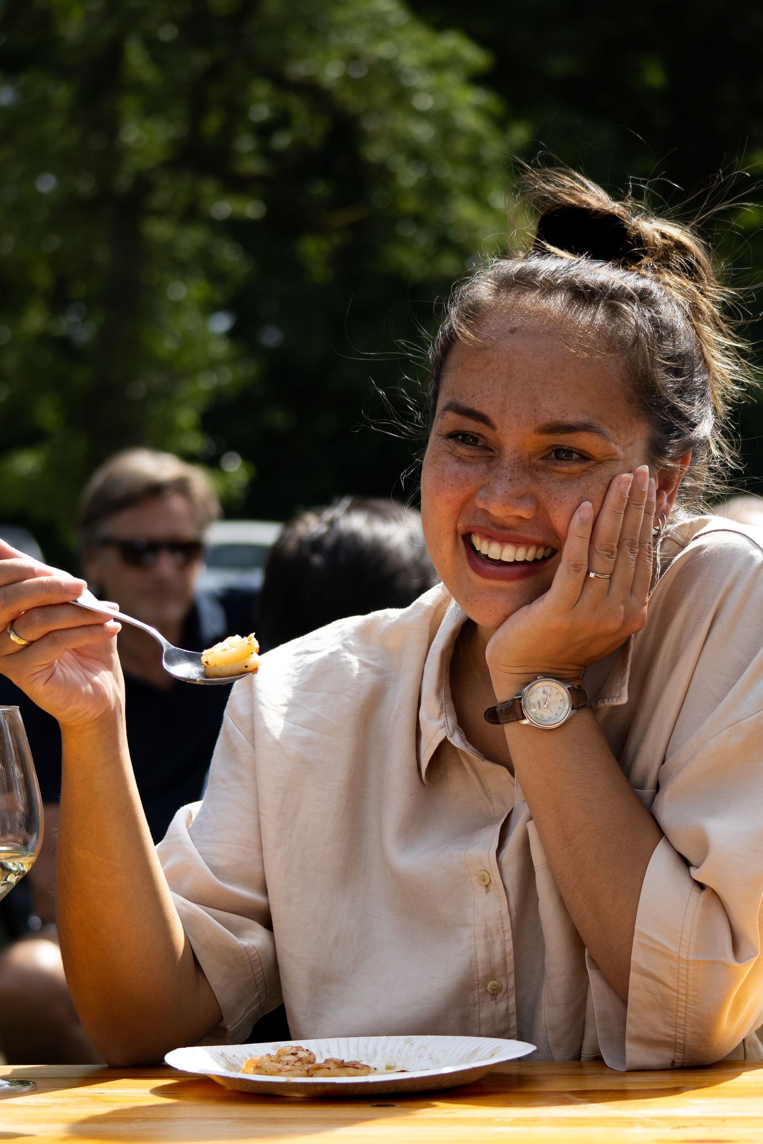 A person in a beige shirt enjoys an outdoor meal on a sunny day while sitting at a wooden table.