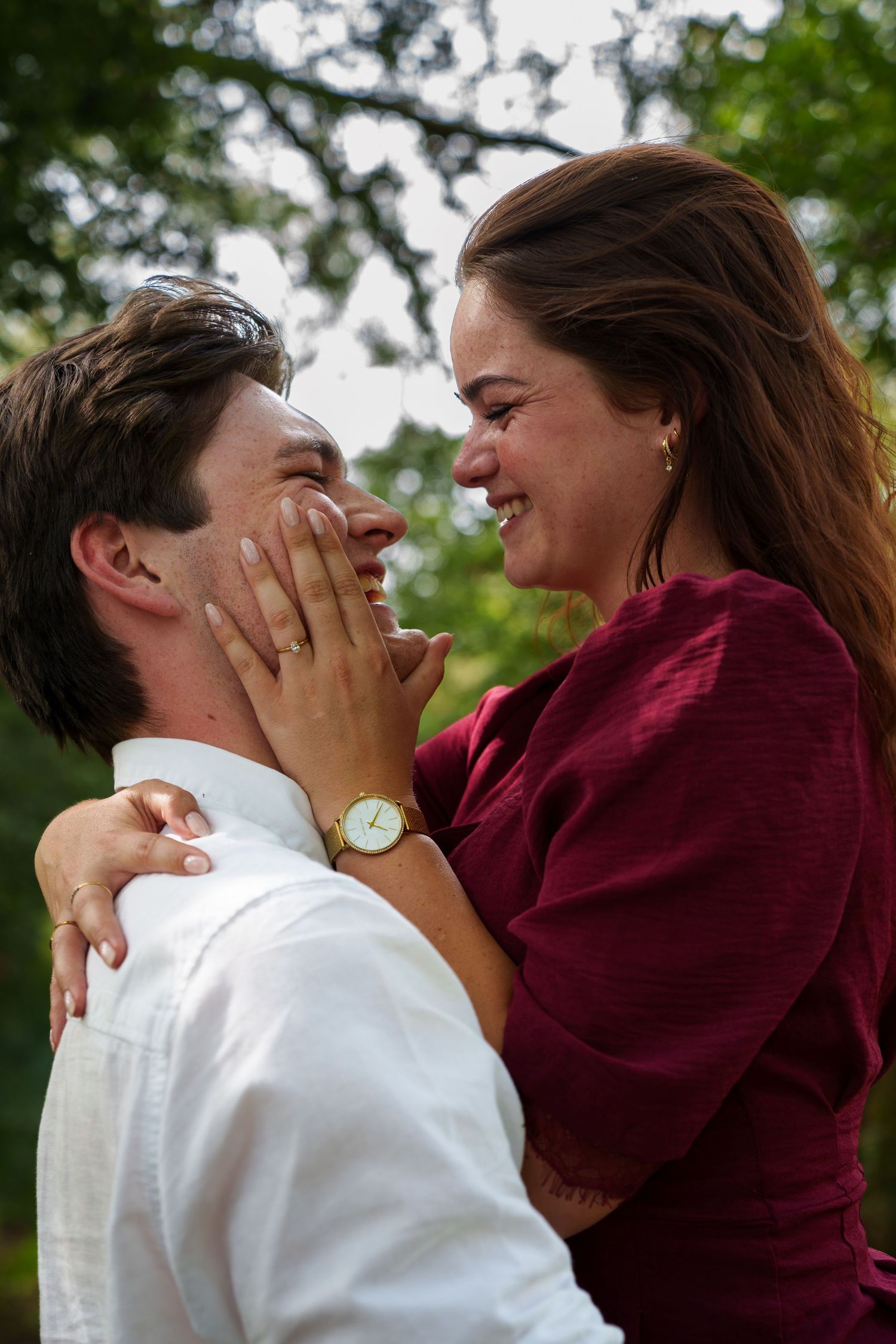 A joyful couple embraces outdoors, woman in burgundy dress covers man's face with her hand, both laughing.