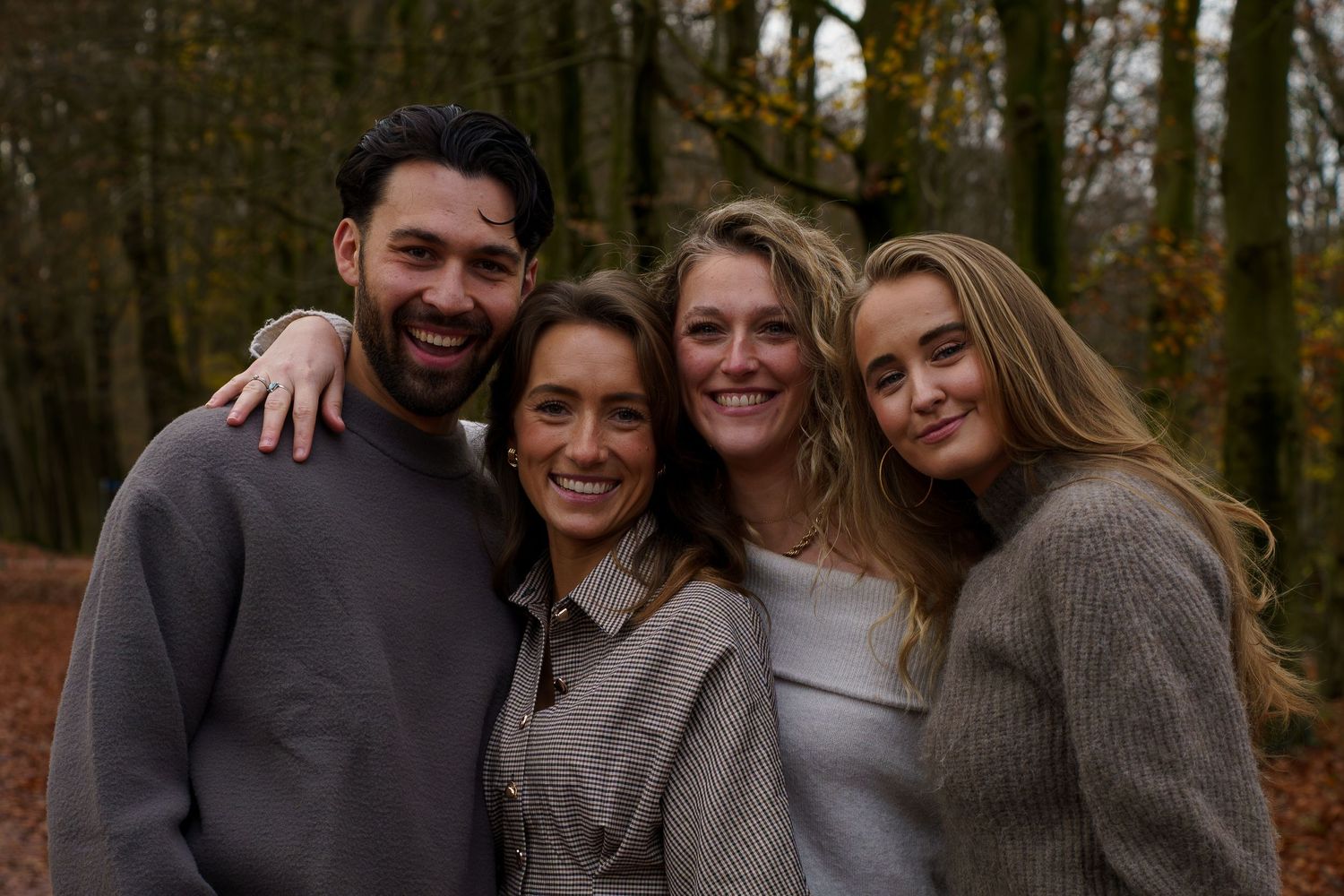 Four smiling friends posing together in an autumn forest with colorful foliage in the background.