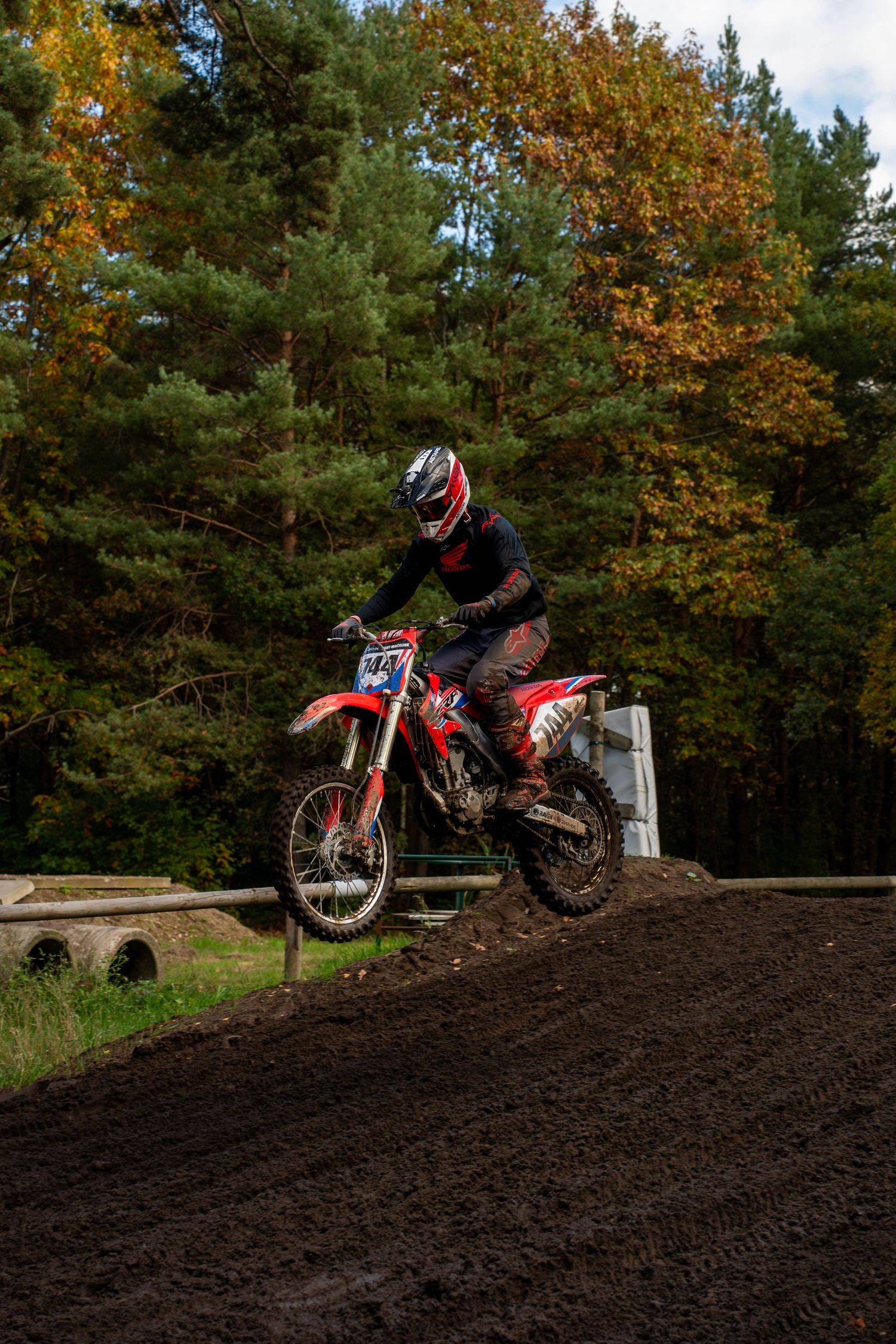 Dirt bike rider navigates a steep dirt mound on a red motorcycle against an autumn forest backdrop.
