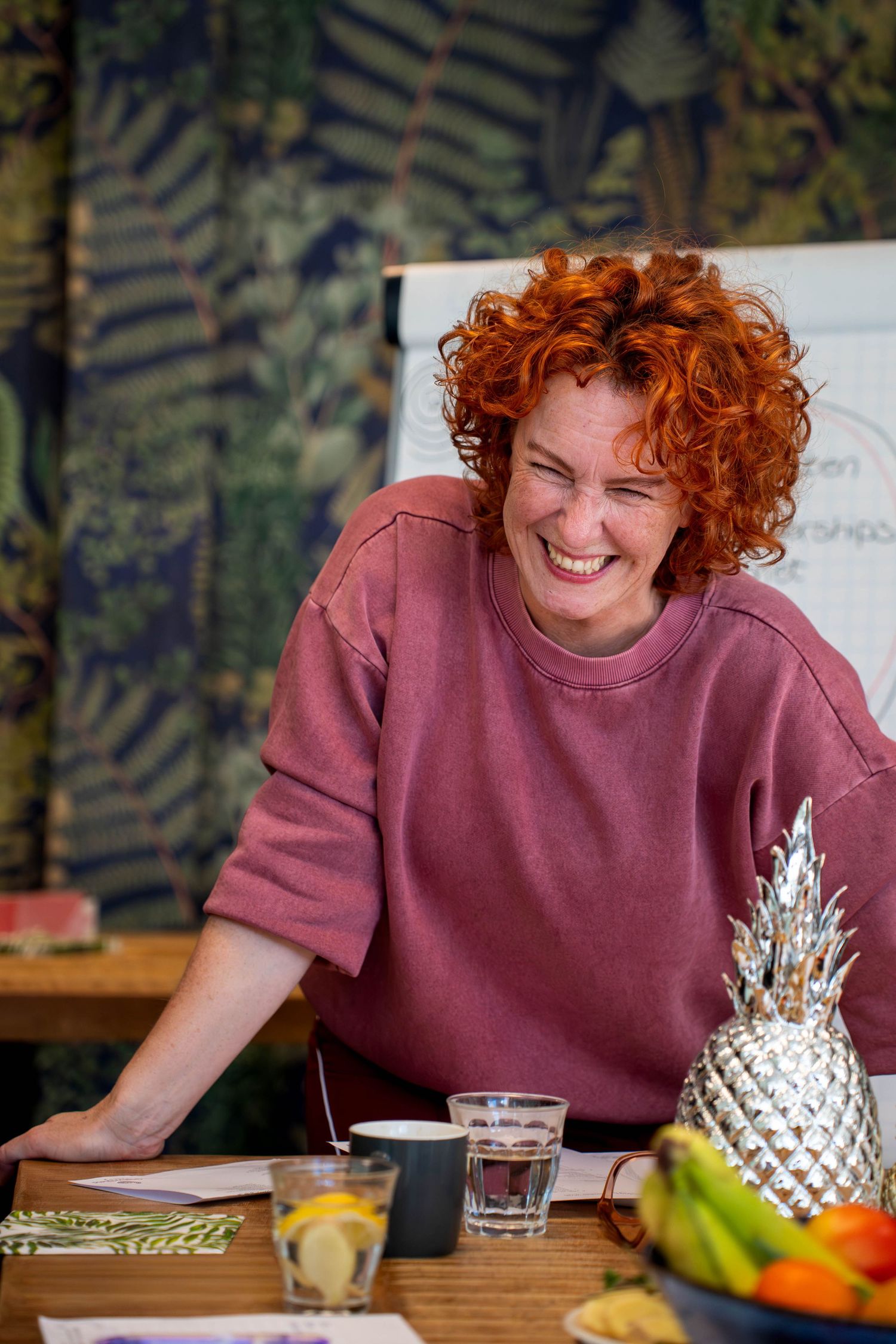 Smiling redhead woman in pink sweatshirt leaning on table with pineapple decor and tropical wallpaper background.