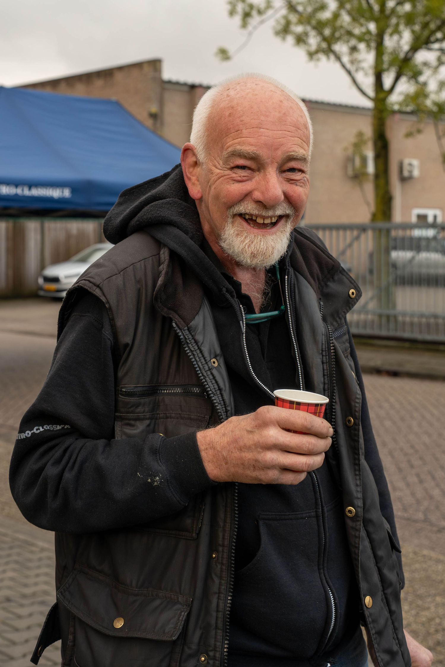 Smiling elderly man with white beard holding a red cup outdoors, wearing a black vest jacket.
