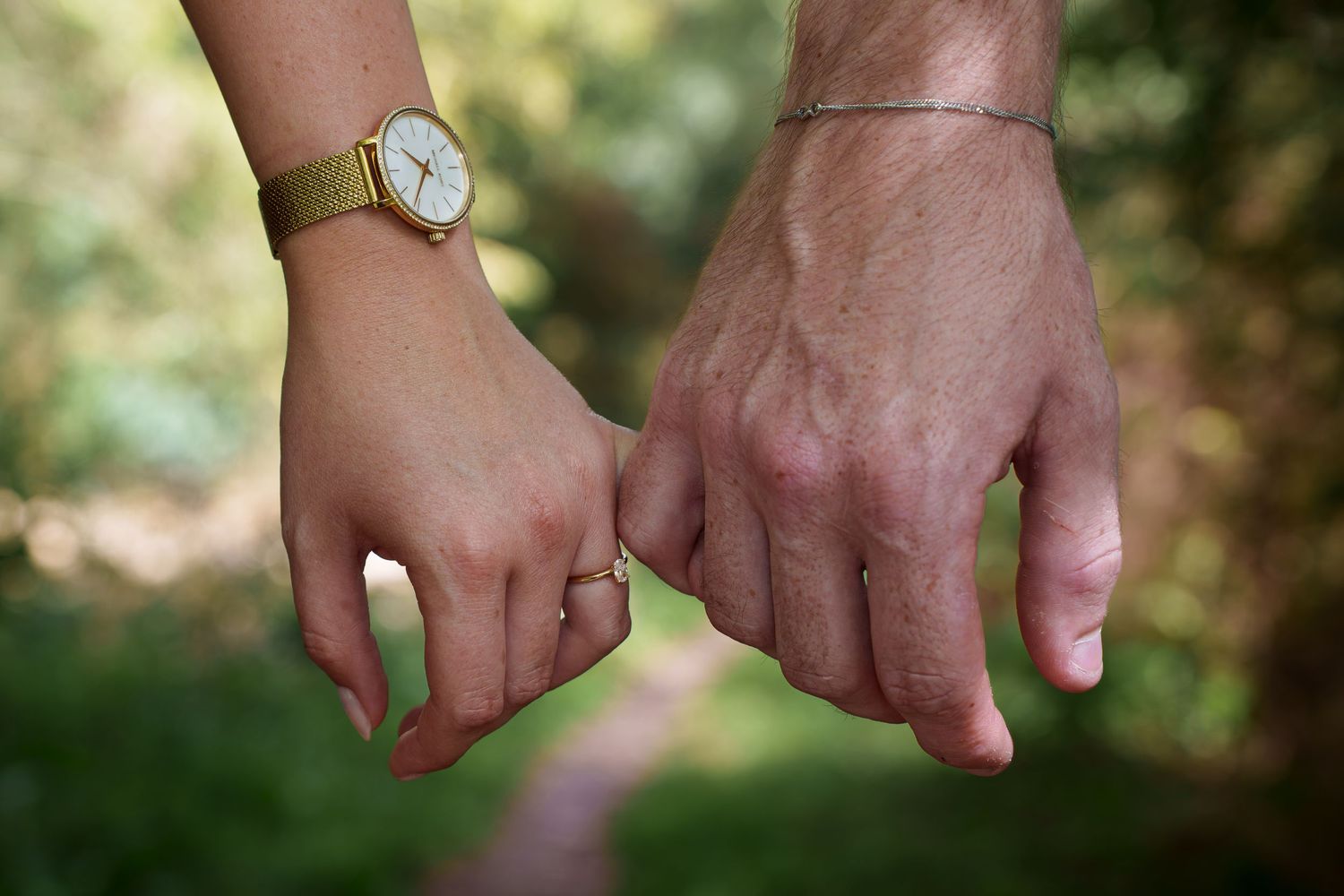 Two hands holding pinkies, one wearing a gold watch and ring, the other a silver bracelet, on a blurred green path.