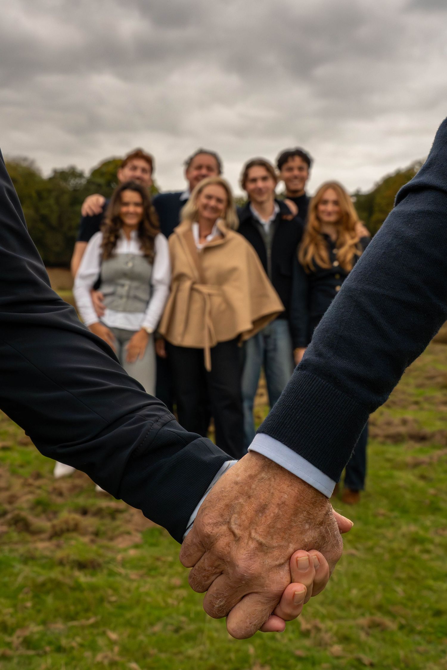 Two people holding hands in foreground with a smiling family group standing in a field under cloudy skies.