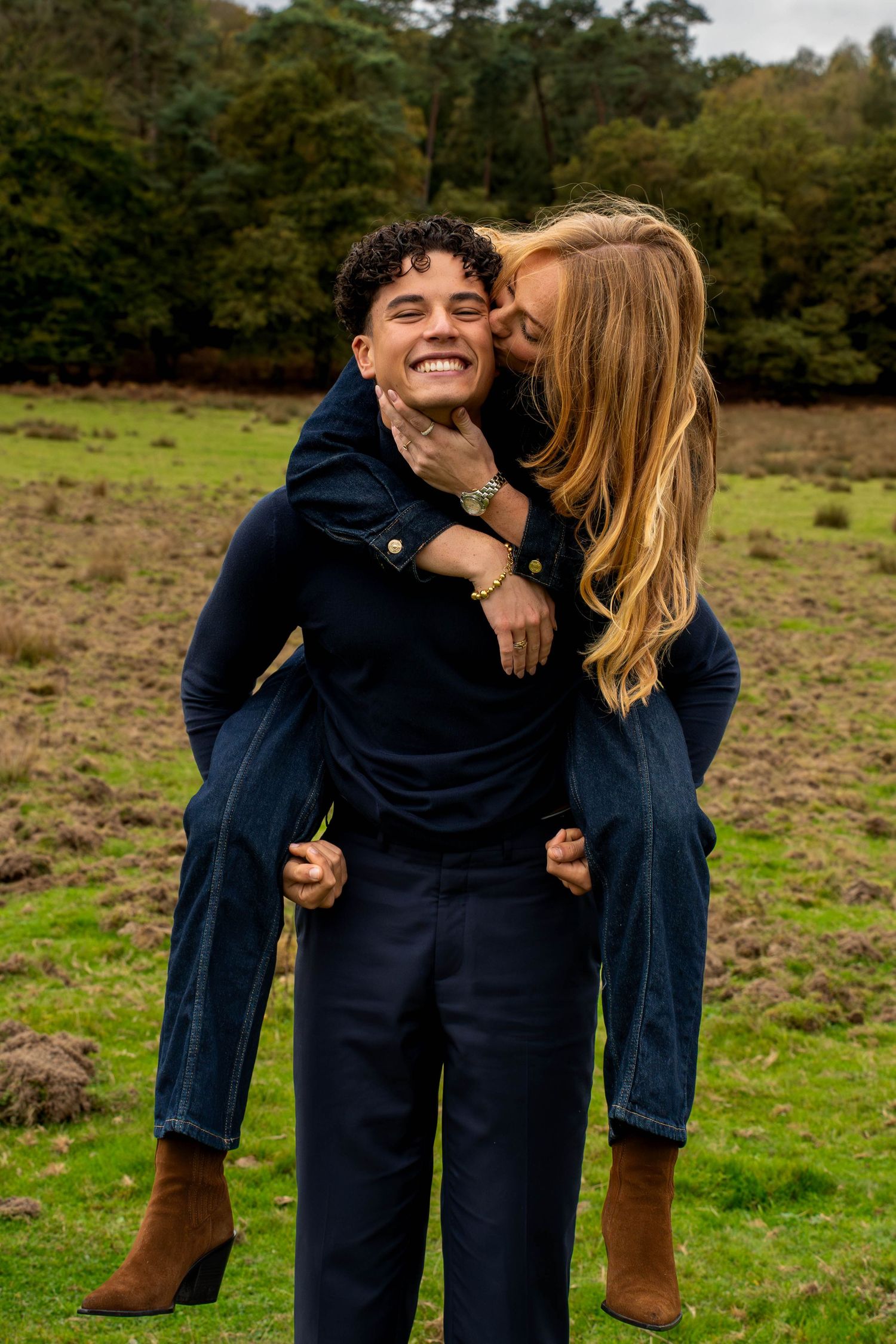 A young man gives a piggyback ride to a smiling blonde woman in a green field, both wearing navy outfits.