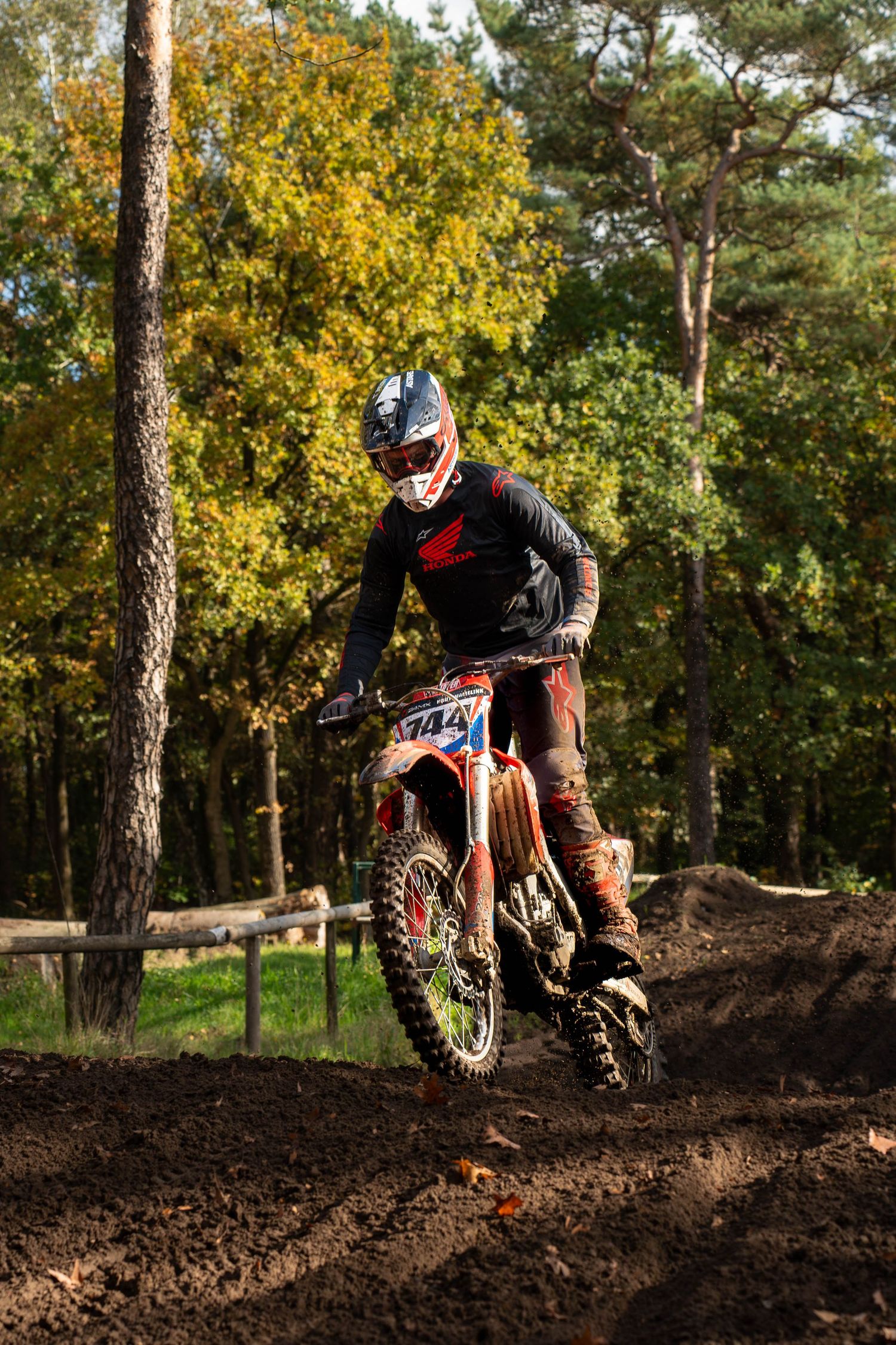 Motocross rider #144 navigating a muddy dirt track on a red bike, surrounded by autumn trees.