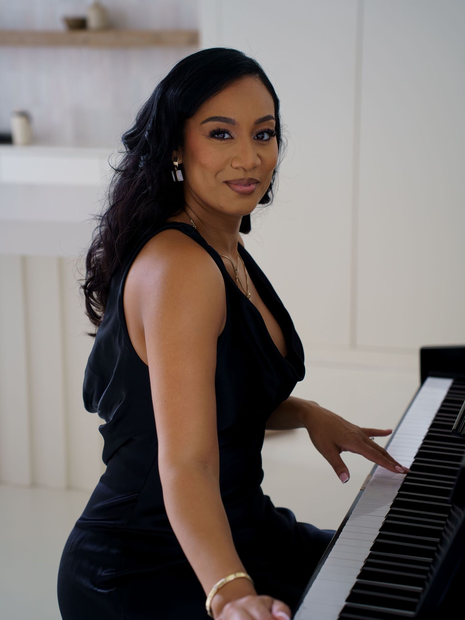 Woman in black dress sitting at piano, smiling over shoulder in bright white room.