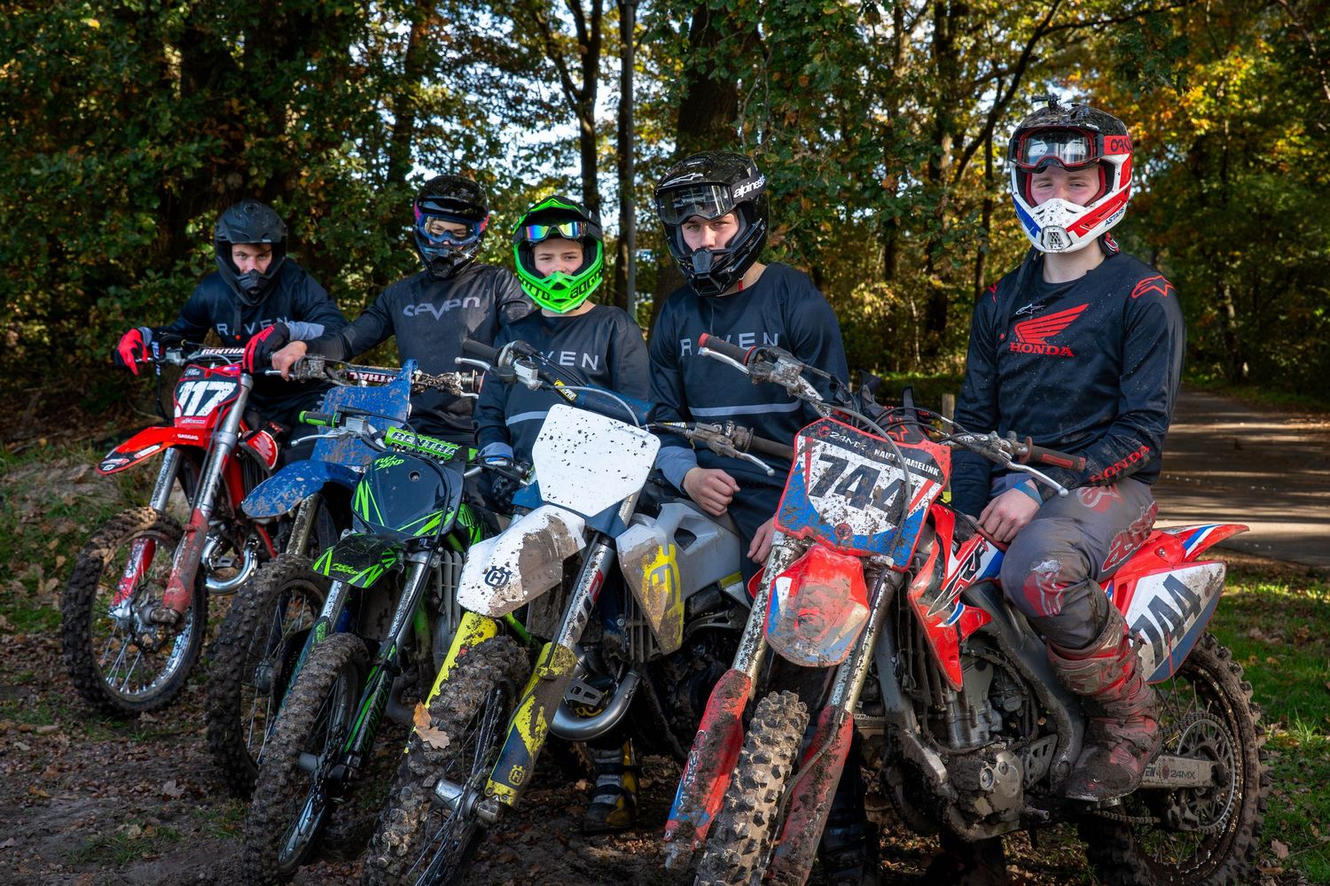 Four motocross riders in gear pose with dirt bikes numbered 17 and 704 on a forest trail.